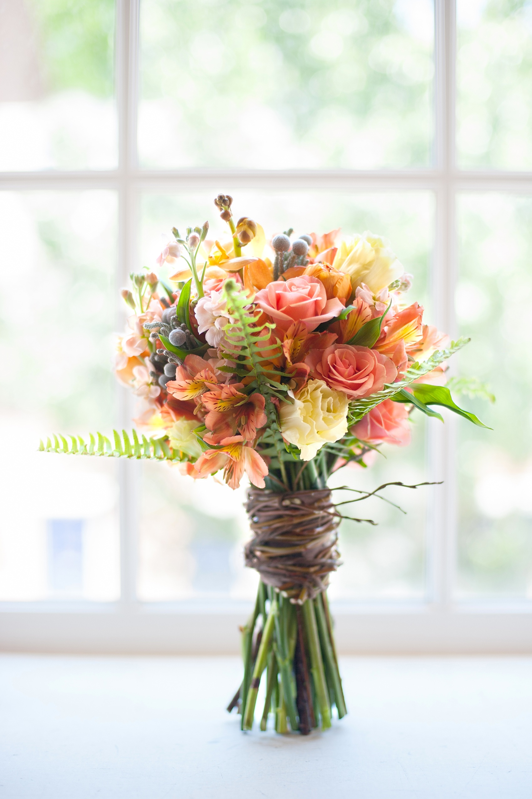 Rustic wedding bouquet tied with willow branches containing green ferns, orange flowers and berries. Photo by Mikkel Paige Photography. 