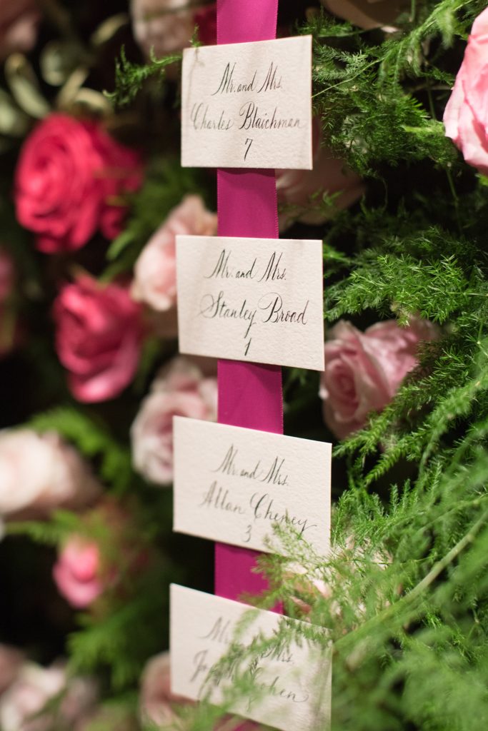 Mikkel Paige Photography wedding photos at The Glasshouses NYC. A flower wall escort card presentation with pink ribbon and calligraphy.