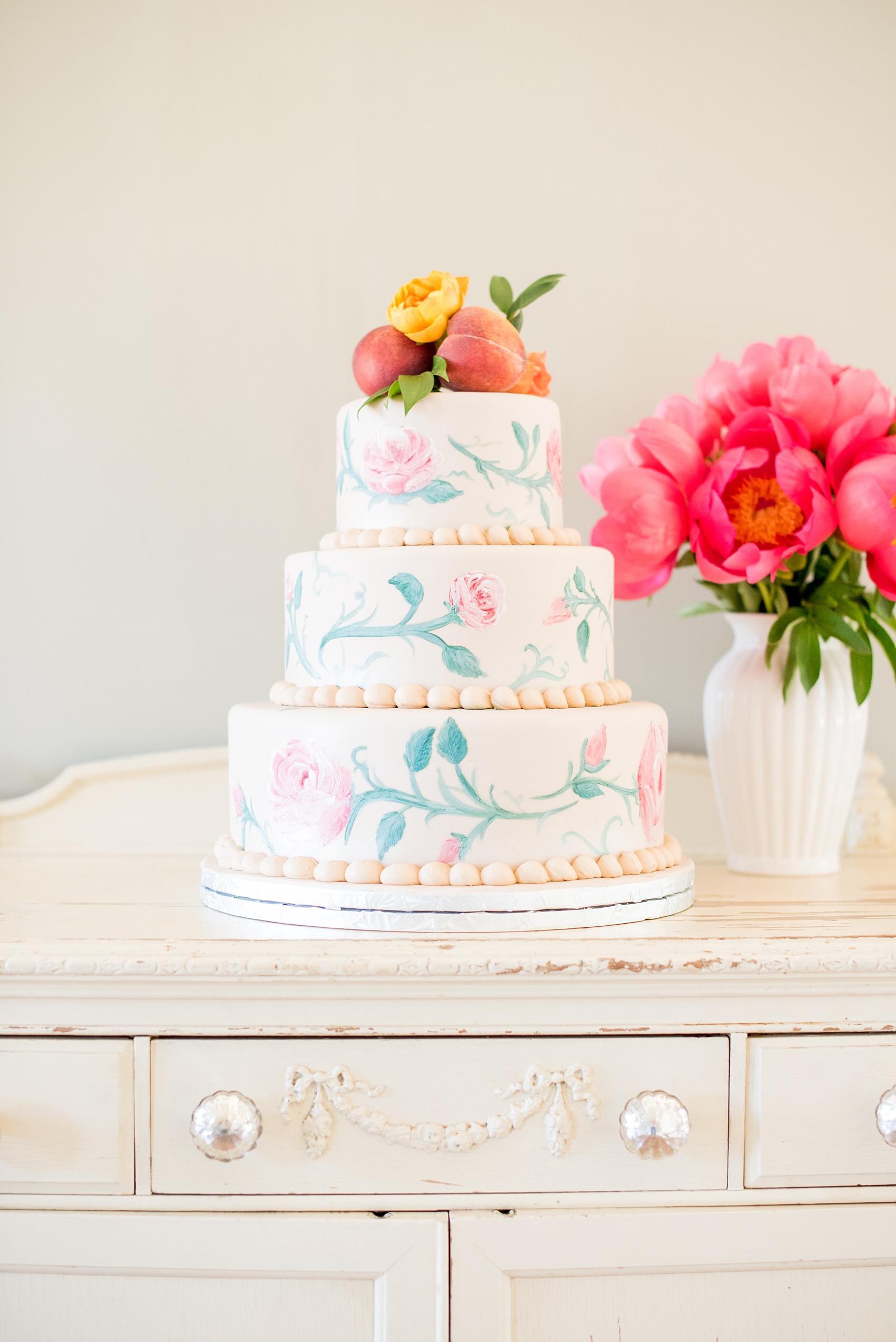 Mikkel Paige Photography photo at The Bradford, NC. Edible art tiered wedding cake in ivory fondant and hand painted pink roses and vines, topped with peaches and yellow ranunculus flower.