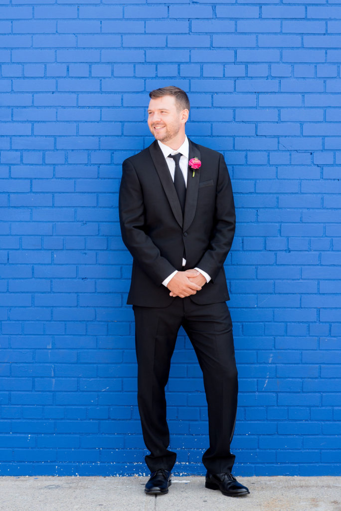 Mikkel Paige Photography photo of Dobbin St Brooklyn wedding. Planning and coordination by Color Pop Events. Tuxedo from The Black Tux for the groom, image against a colorful blue wall.