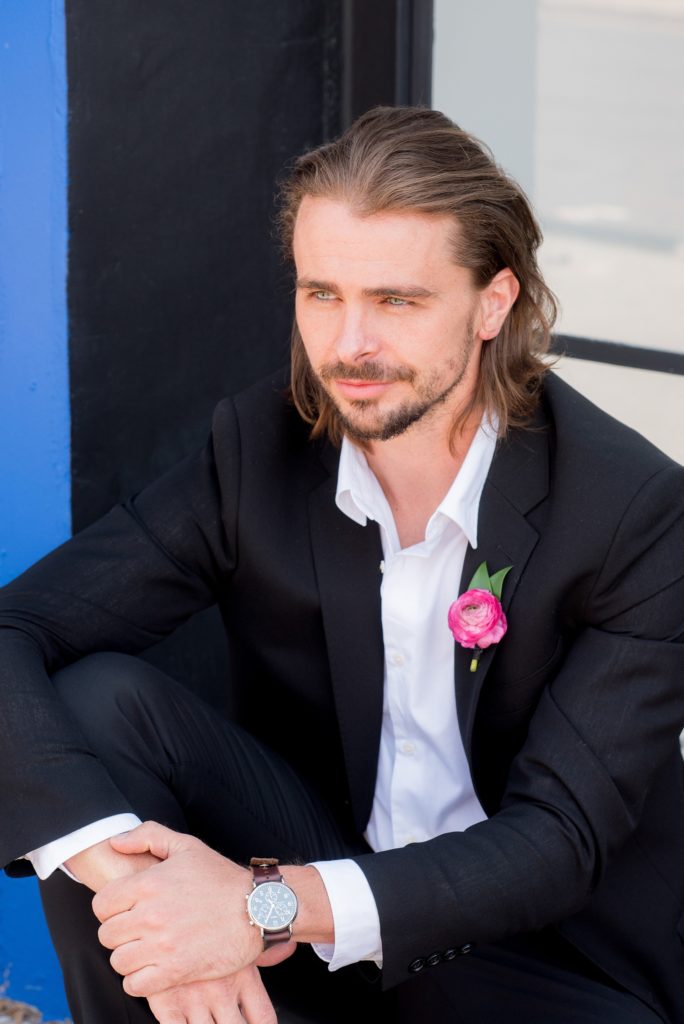 Mikkel Paige Photography photo of Dobbin St Brooklyn wedding. Planning and coordination by Color Pop Events. Long hair groomsmen in a classic black tuxedo from The Black Tux, image against a colorful blue wall in Williamsburg.