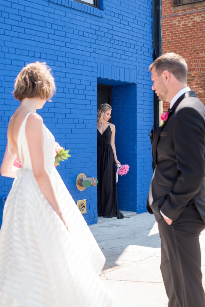 Mikkel Paige Photography photo of Dobbin St Brooklyn wedding. Planning and coordination by Color Pop Events. Tuxedo from The Black Tux for the groom, image against a colorful blue wall. Bride in a Hayley Paige white striped gown and bridesmaid in a black dress.