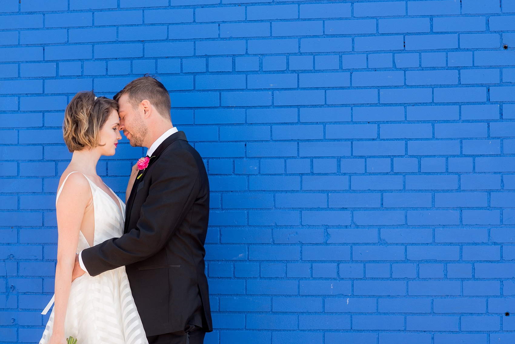 Mikkel Paige Photography photo of Dobbin St Brooklyn wedding. Planning and coordination by Color Pop Events. Tuxedo from The Black Tux for the groom, image against a colorful blue wall. Bride in a Hayley Paige white striped gown.