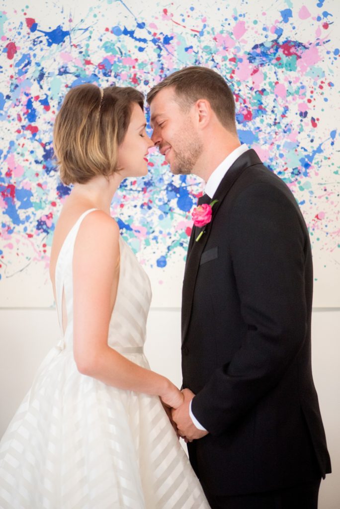 Mikkel Paige Photography photo of Dobbin St Brooklyn wedding. Planning and coordination by Color Pop Events. Ceremony setup with clear chiavari chairs and custom art splatter paint backdrop behind the bride and groom's first kiss.