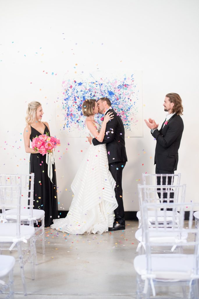 Mikkel Paige Photography photo of Dobbin St Brooklyn wedding. Planning and coordination by Color Pop Events. Ceremony confetti toss with clear chiavari chairs and custom art splatter paint backdrop for the bride and groom's first kiss.