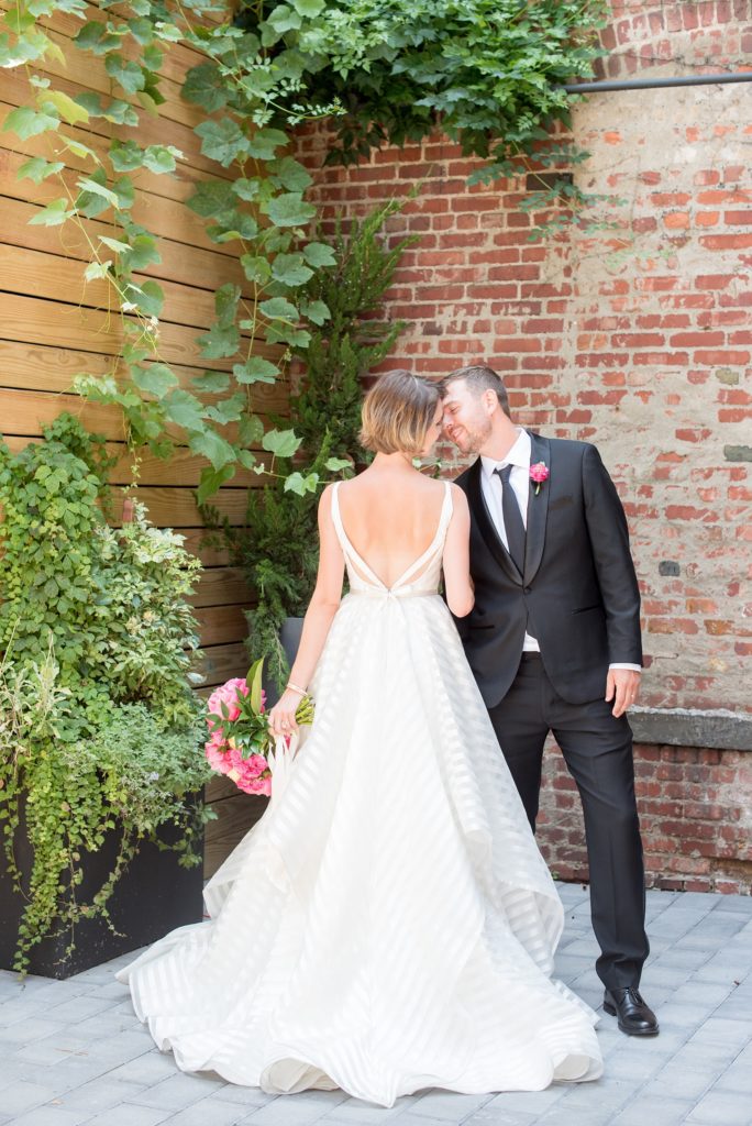 Mikkel Paige Photography photo of Dobbin St Brooklyn wedding. Planning and coordination by Color Pop Events. Bride in a white striped Hayley Paige gown and groom in classic Tuxedo by The Black Tux. Outdoor patio image against greenery and rustic brick.