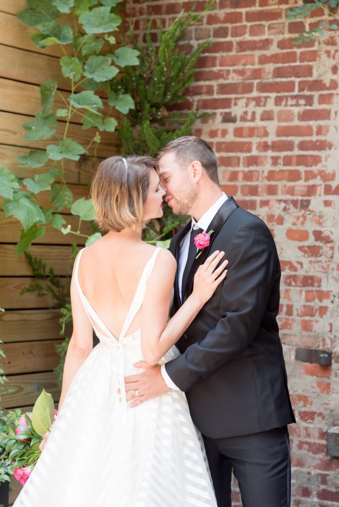 Mikkel Paige Photography photo of Dobbin St Brooklyn wedding. Planning and coordination by Color Pop Events. Bride in a white striped Hayley Paige gown and groom in classic Tuxedo by The Black Tux. Outdoor patio image against greenery and rustic brick.