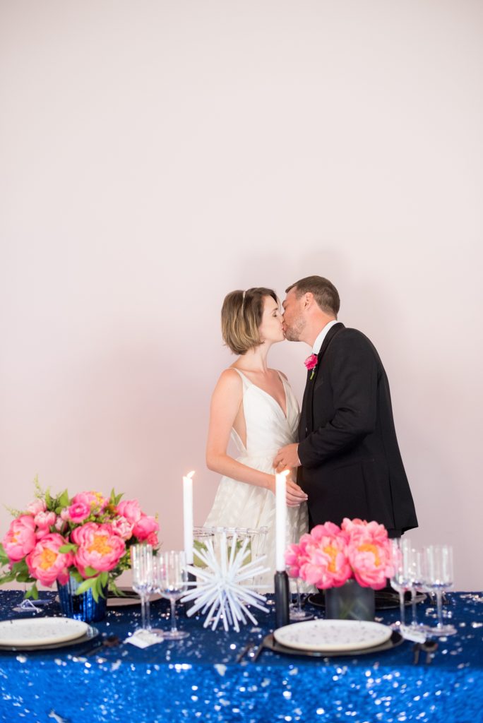 Mikkel Paige Photography photo of Dobbin St Brooklyn wedding. Planning and coordination by Color Pop Events. Reception table with blue sequins, pink peony centerpieces and clear chiavari chairs against a mauve wall with the bride and groom.