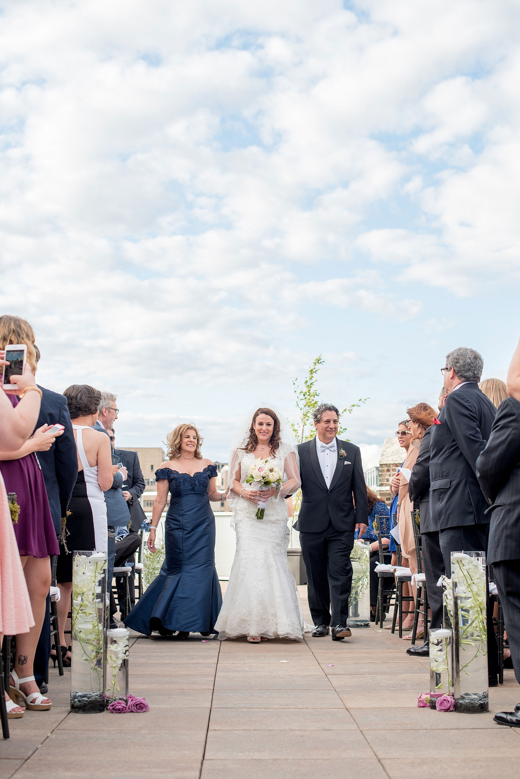 Mikkel Paige Photography photos of a NYC wedding at Tribeca Rooftop. An image of the bride and her parents walking down the aisle. She wore a lace cap sleeve Anita Graham gown.