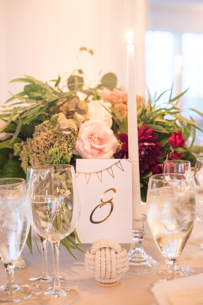 Mikkel Paige Photography picture of a Bay Head Yacht Club nautical wedding. Gold table numbers were help in white rope spheres next to gold flatware with fall colored floral centerpieces and tapered candles.