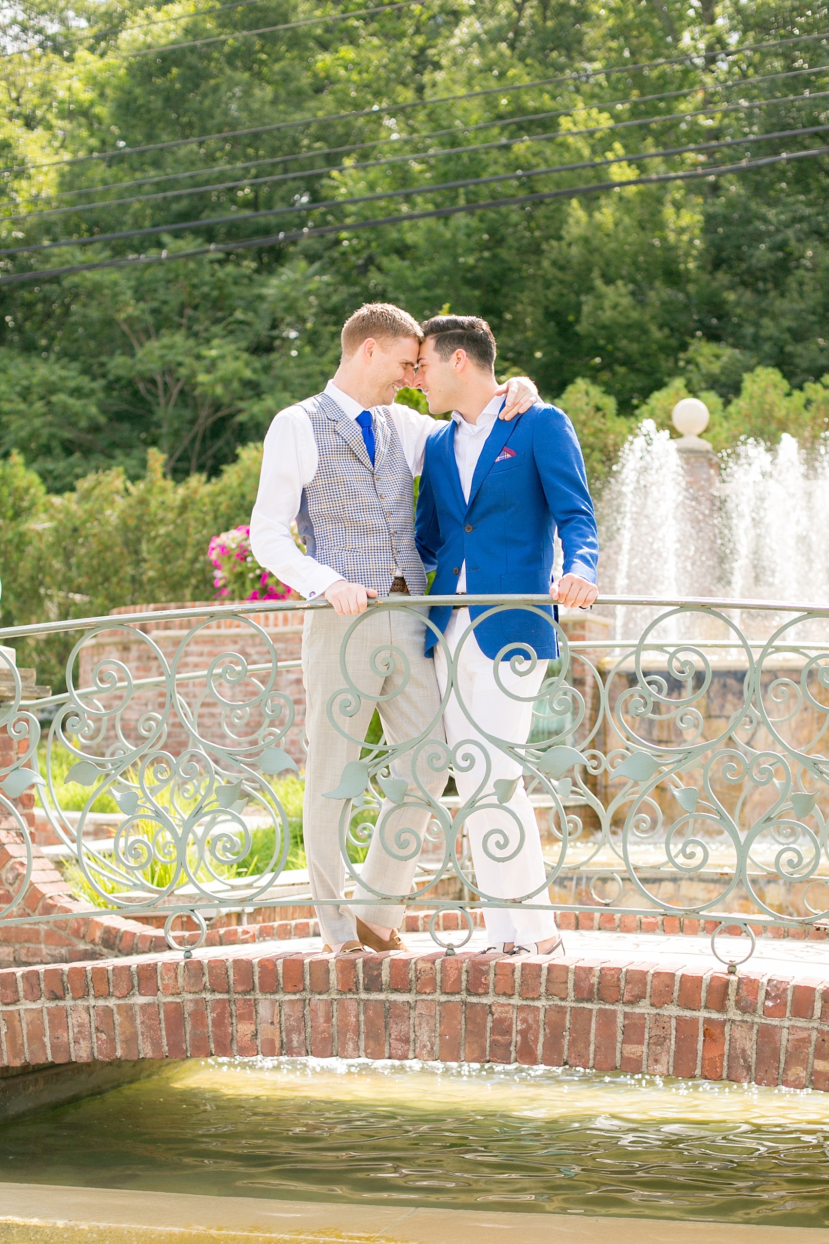 Mikkel Paige Photography photos of a summer, daytime gay wedding. An image of the grooms in a vest and blue suit on a picturesque bridge in front of a fountain at The Manor, in West Orange New Jersey.