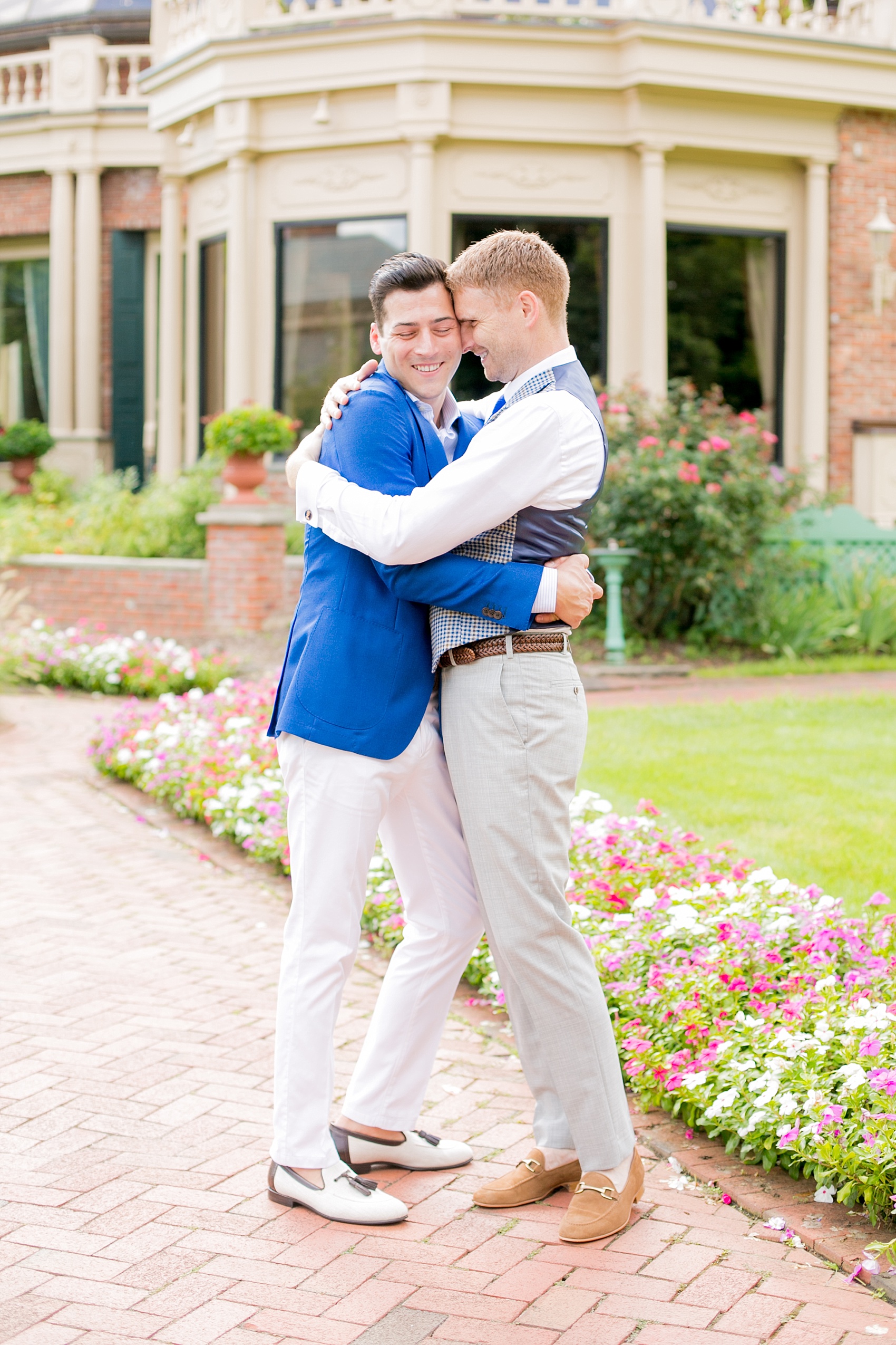 Mikkel Paige Photography photos of a summer, daytime gay wedding. A romantic, playful image of the grooms hugging in a vest and blue suit in front of picturesque landscaping at The Manor, in West Orange New Jersey.