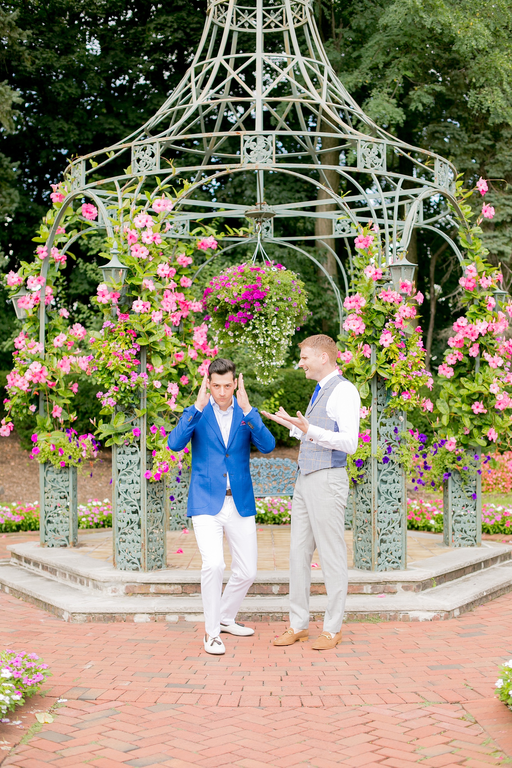 Mikkel Paige Photography photos of a summer, daytime gay wedding. An image of the grooms in a vest and blue suit in front of a trellis dome with pink and green landscaping at The Manor, in West Orange New Jersey.