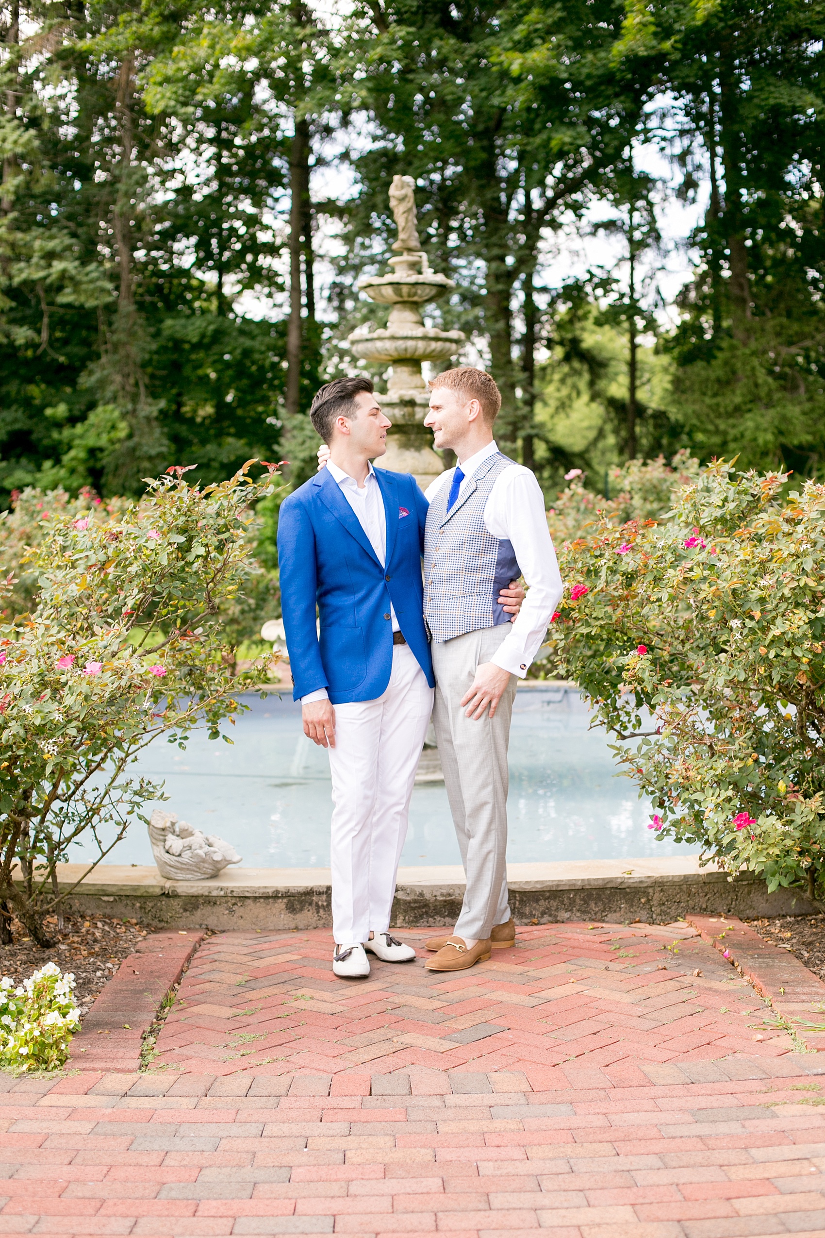 Mikkel Paige Photography photos of a summer, daytime gay wedding. An image of the grooms in a vest and blue suit in front of a fountain and pool, with rose bushes at The Manor, in West Orange New Jersey.