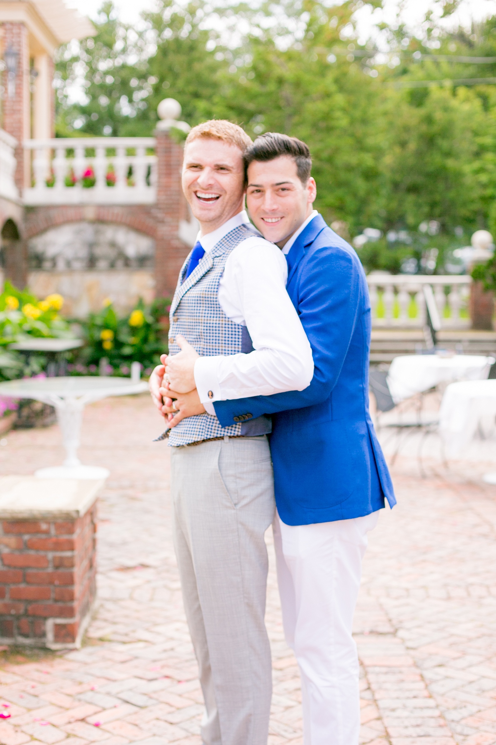 Mikkel Paige Photography photos of a summer, daytime gay wedding. A romantic, playful image of the grooms hugging in a vest and blue suit in front of picturesque landscaping at The Manor, in West Orange New Jersey.