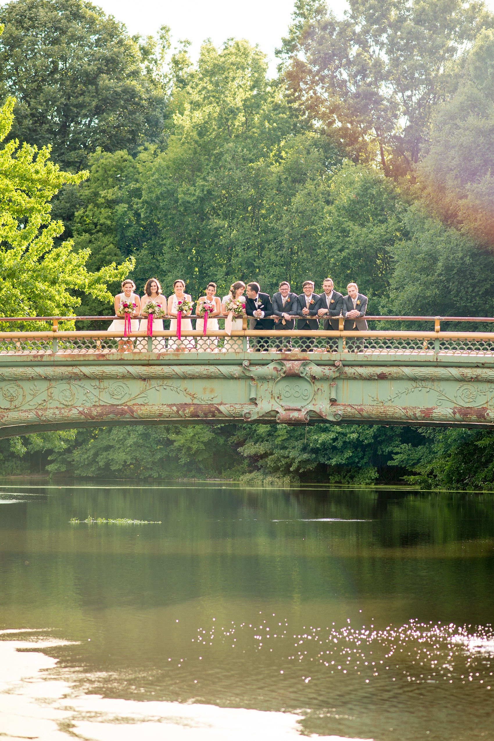 Mikkel Paige Photography photos of a wedding at Prospect Park Boathouse in Brooklyn, NY. Bridal party photo on a bridge overlooking water with an all-white bridal party and groomsmen in grey suits.