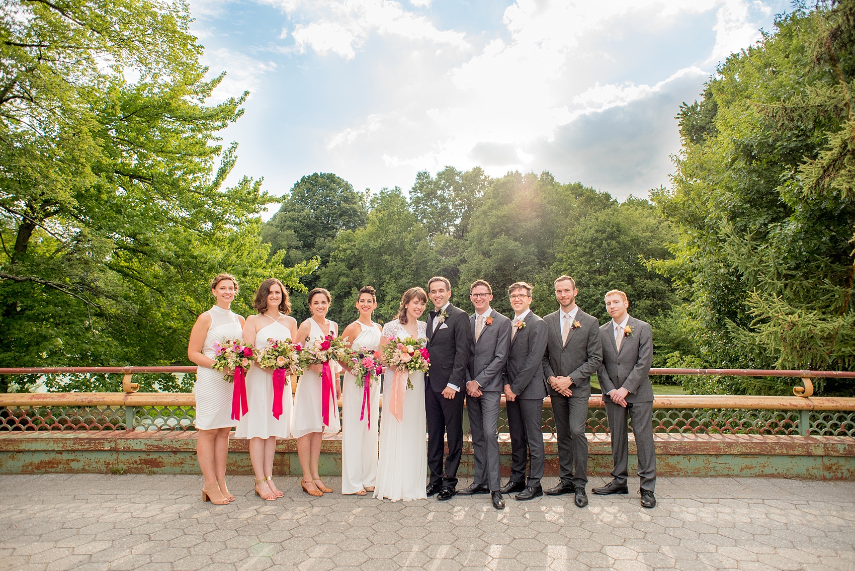 Mikkel Paige Photography photos of a wedding at Prospect Park Boathouse in Brooklyn, NY. Bridal party photo on a bridge overlooking water with an all-white bridal party and groomsmen in grey suits.