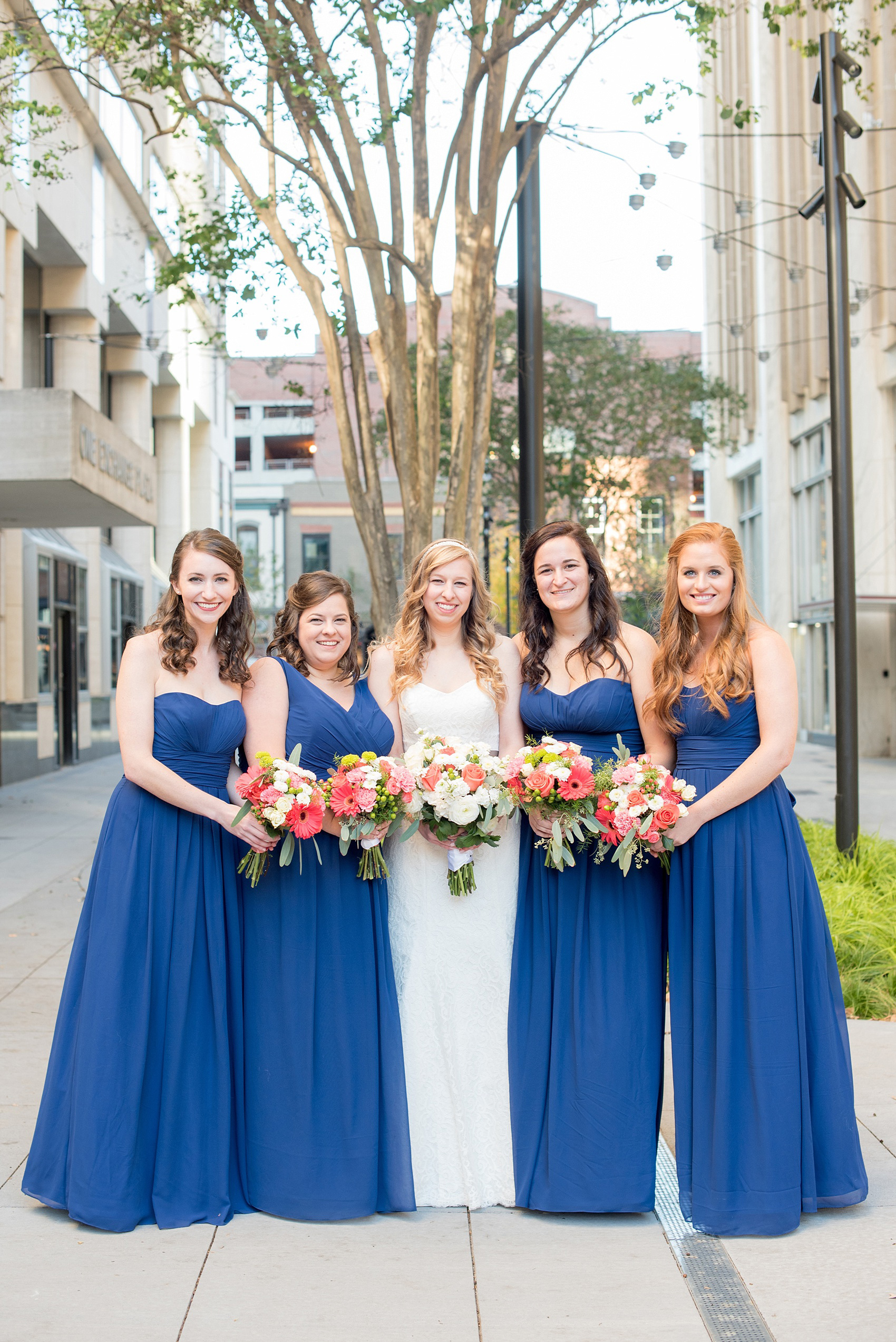 Mikkel Paige Photography photos from a wedding at The Stockroom at 230. A picture of the bride and her bridesmaids in cobalt blue in downtown Raleigh with coral and white bouquets.
