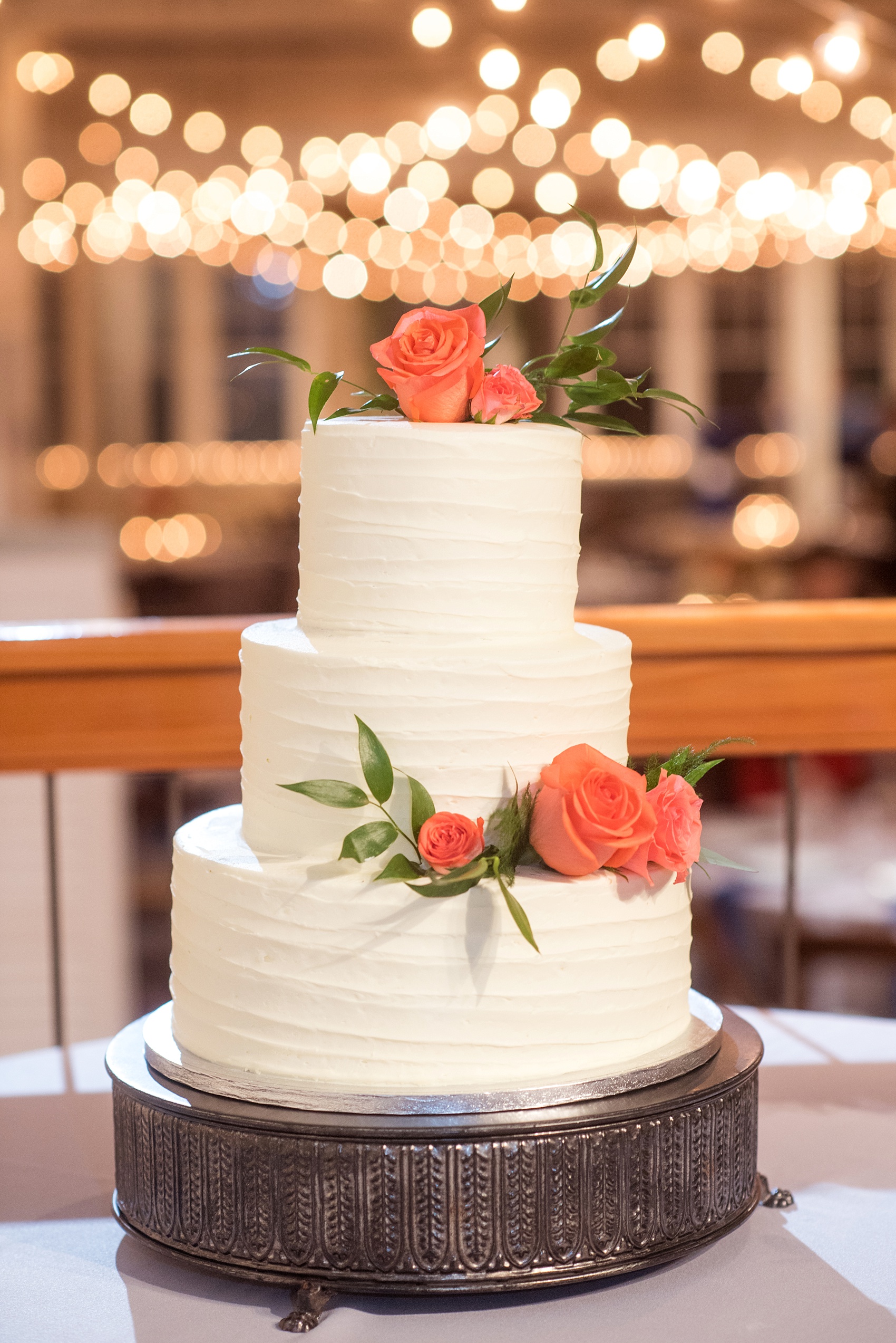 Mikkel Paige Photography photos from a wedding reception at The Stockroom at 230 in Raleigh. A picture of the white tiered buttercream cake adorned with coral roses.