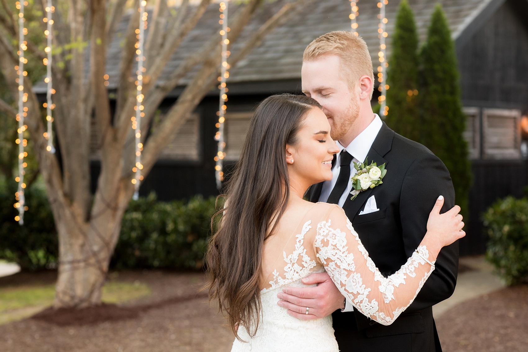 The Sutherland Wedding Photos by Mikkel Paige Photography. The bride in a long-sleeve lace Pronovias gown and groom in black suit, privately share a kiss in front of hanging twinkle lights in this creative picture.