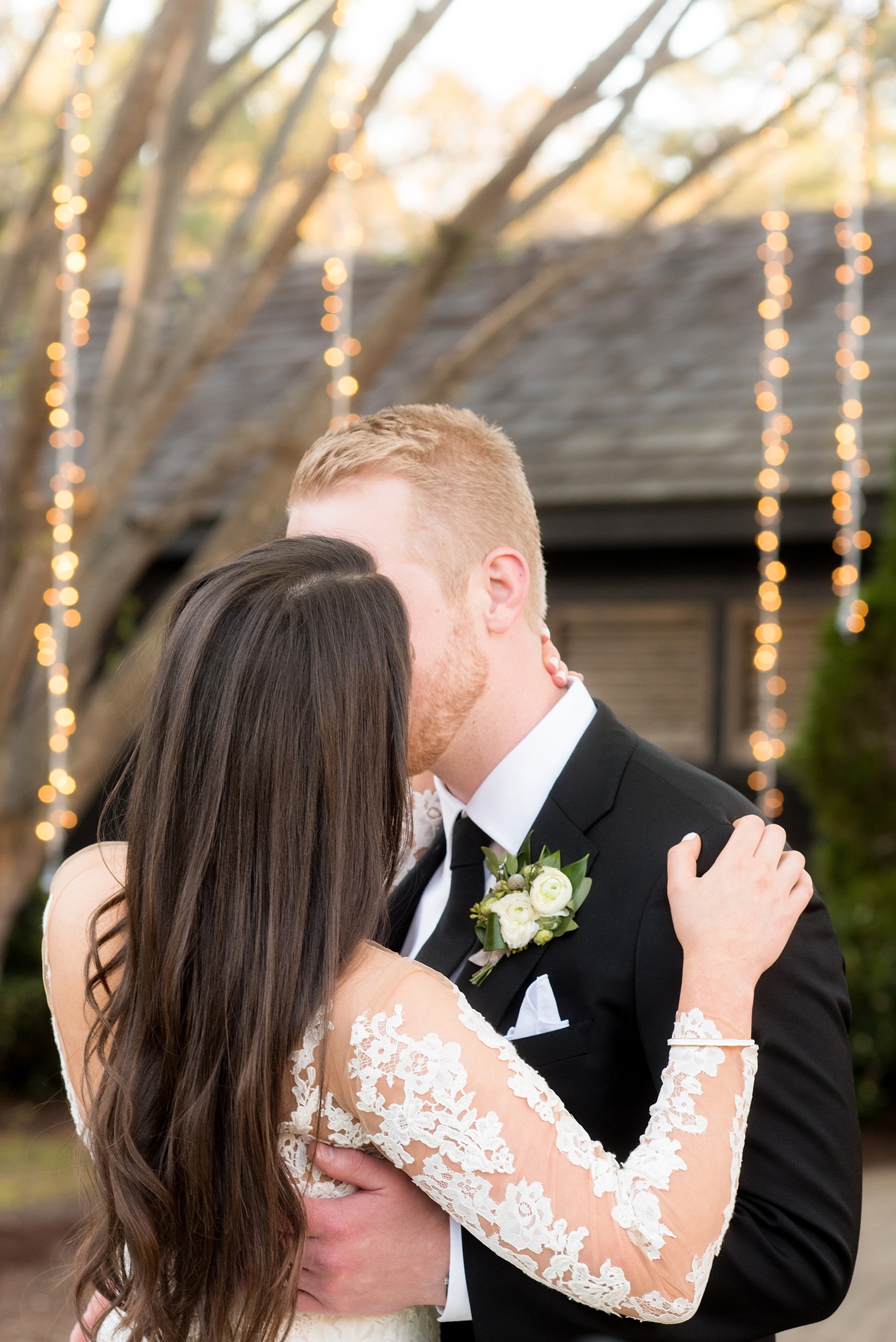 The Sutherland Wedding Photos by Mikkel Paige Photography. The bride in a long-sleeve lace Pronovias gown and groom in black suit, privately share a kiss in front of hanging twinkle lights in this creative picture.