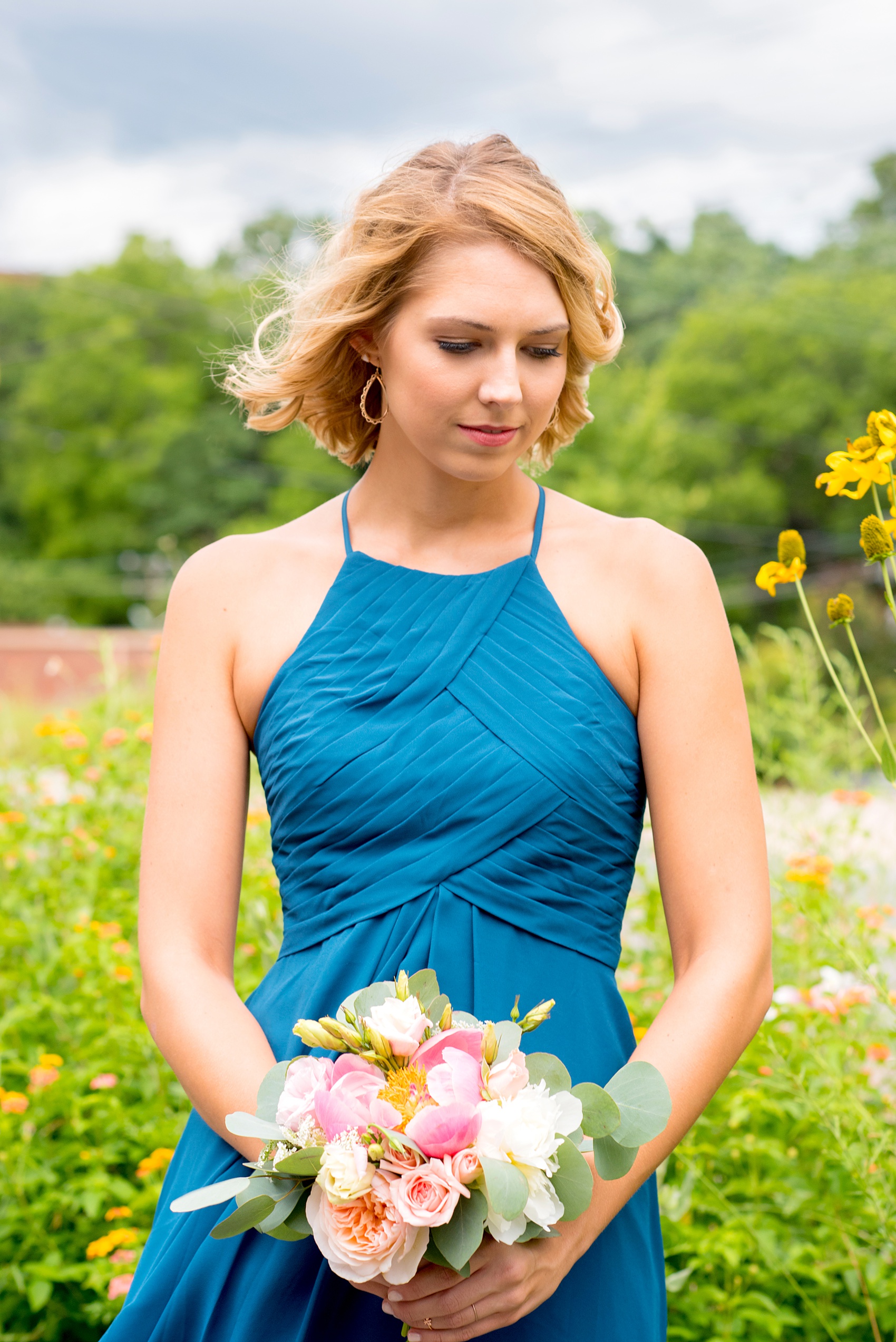 Mikkel Paige Photography photos from a wedding in Durham, North Carolina. Picture of the maid of honor with yellow flowers behind her, in a teal halter top dress, holding a bouquet of peonies and eucalyptus.