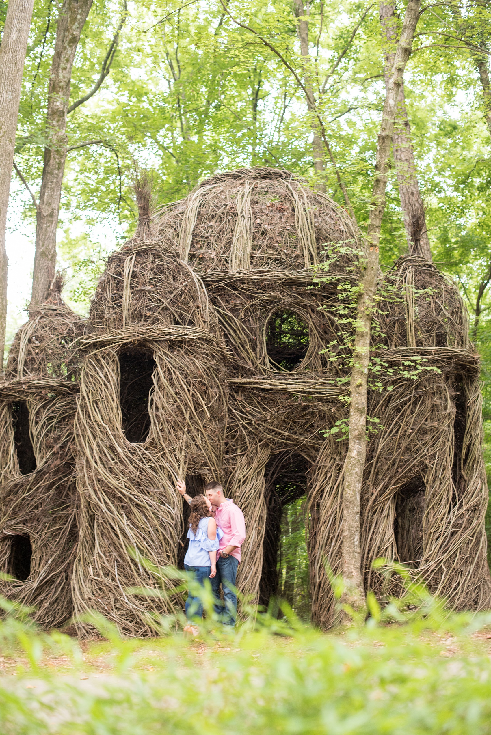 Mikkel Paige Photography photos of an engagement session at Eno Riverwalk in Hillsborough, North Carolina. Picture of the bride and groom kissing with a Stick Art branch sculpture in the background.