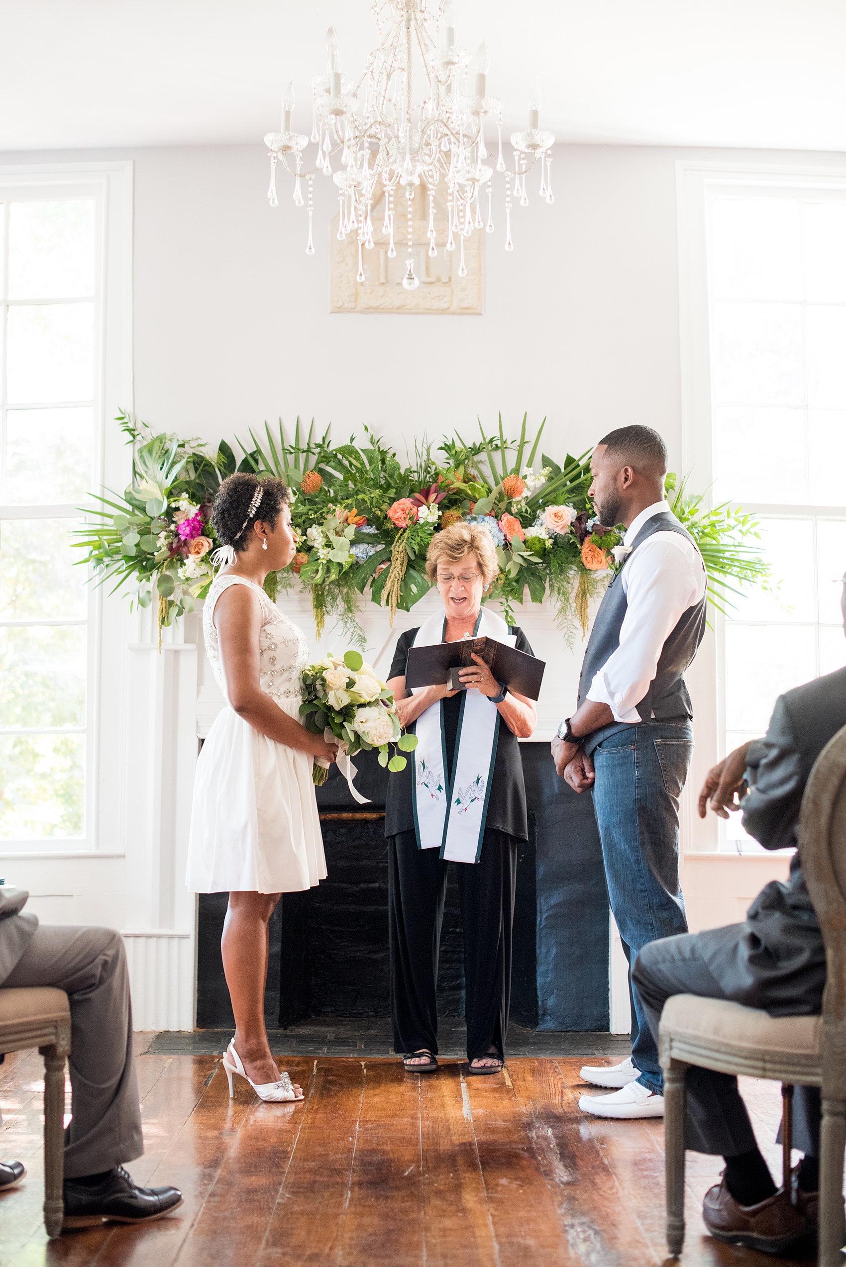 Mikkel Paige Photography pictures of a wedding at Leslie-Alford Mim's House in North Carolina for a Mad Dash Weddings event. Photo of the small elopement ceremony with the bride and groom inside the historic home with a tropical floral arrangement above the fireplace.