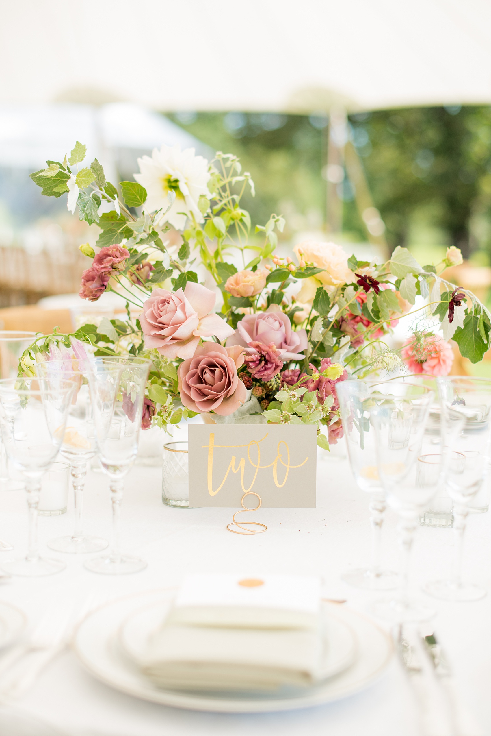 Mikkel Paige Photography photos from a Southwood Estate Wedding in Germantown, New York in the Hudson Valley. Picture of the centerpiece on a table with a grey card and gold metallic script table number and flowers including mauve, dusty rose, burgundy lavender, and pink roses, dahlias, spray roses, peaches and berries.