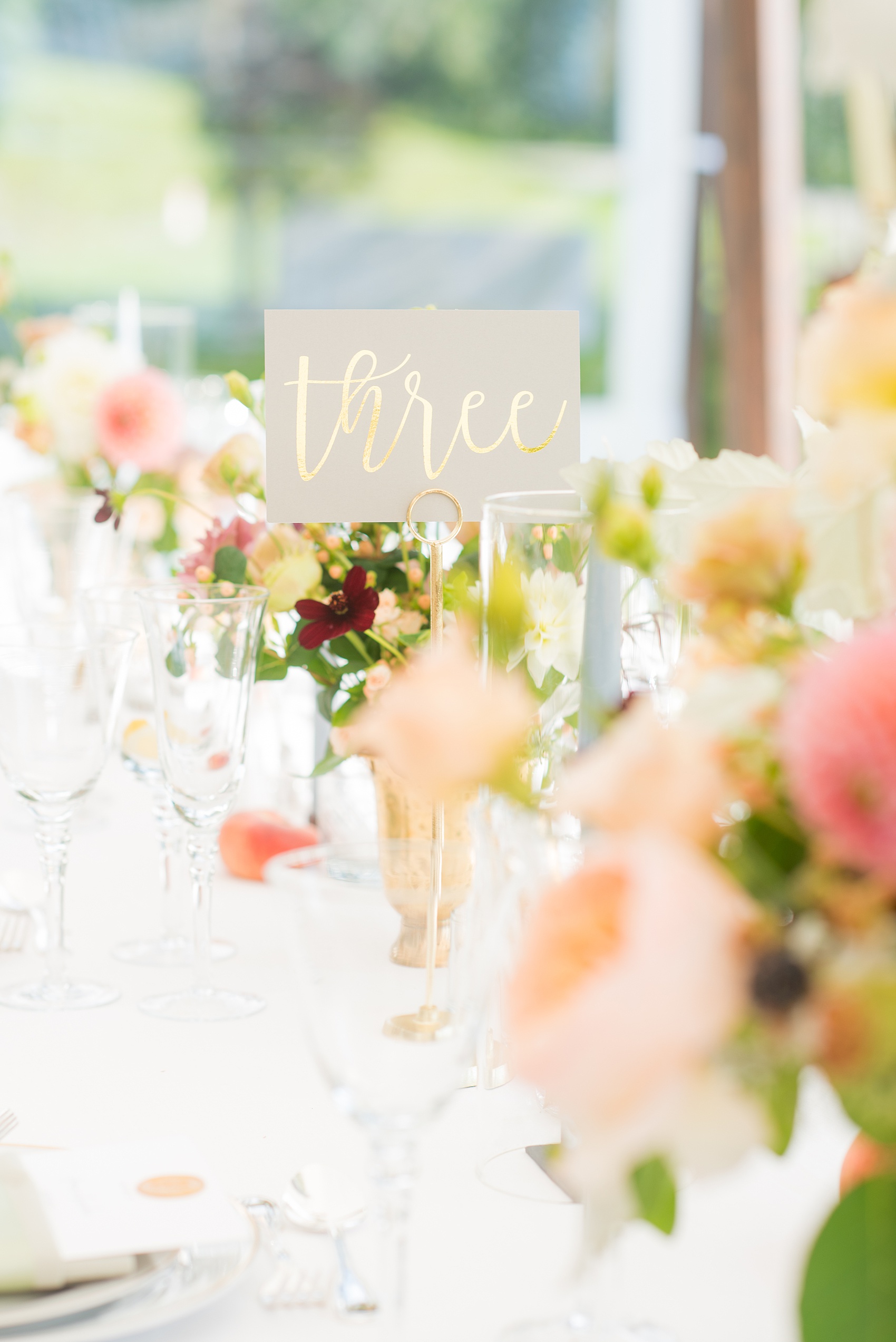 Mikkel Paige Photography photos from a Southwood Estate Wedding in Germantown, New York in the Hudson Valley. Picture of the table numbers with a grey card and gold metallic script and colorful floral centerpieces.