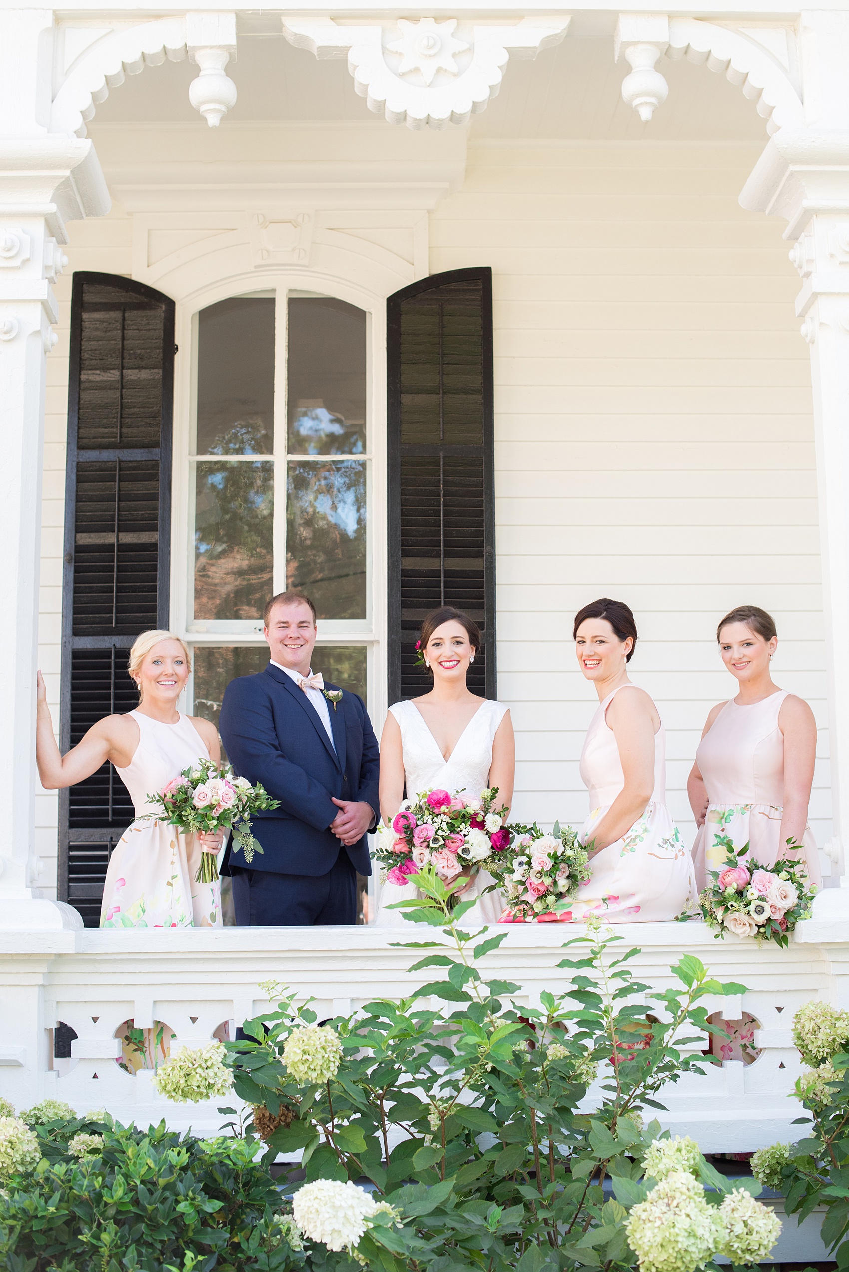 Mikkel Paige Photography pictures from a wedding at Merrimon-Wynne House in Raleigh, NC. Photo of the bridal party on the porch of the historic home. The bridesmaids wore pink dresses with a colorful floral pattern.