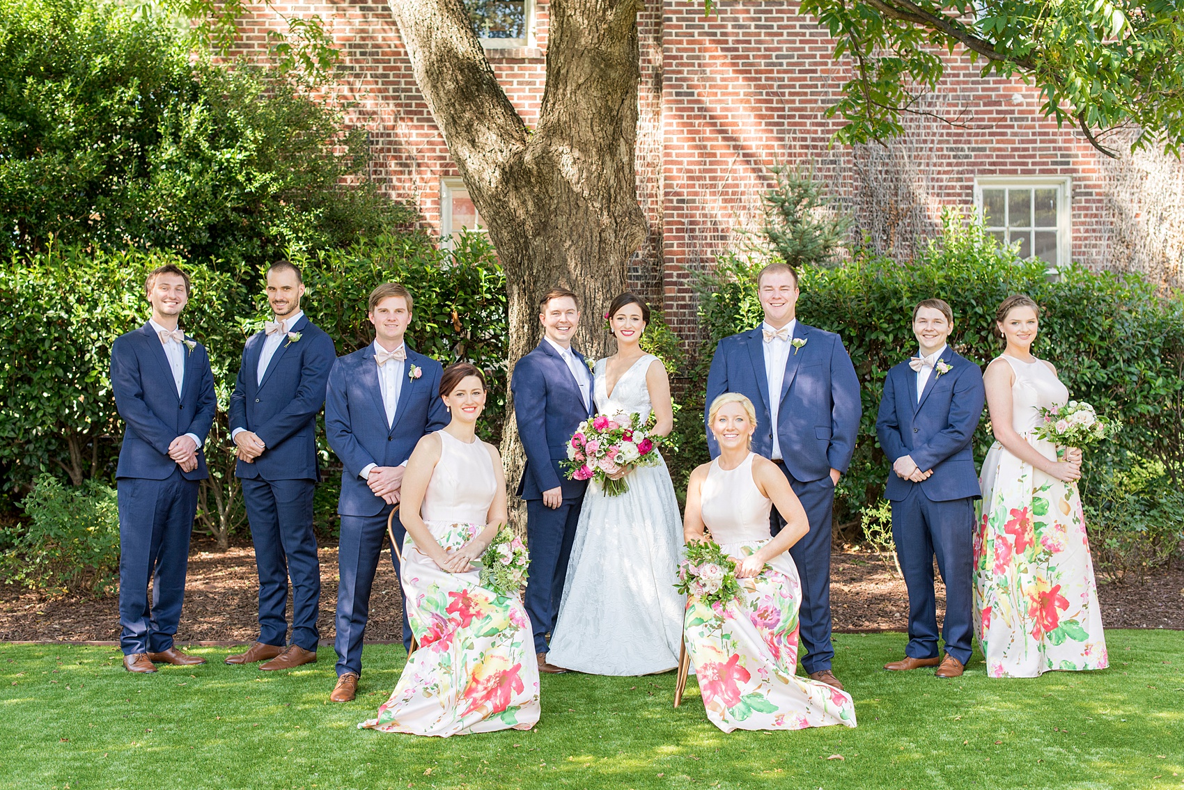 Mikkel Paige Photography pictures from a wedding at Merrimon-Wynne House in Raleigh, NC. Creative photo of the bridal party on the lawn of the historic home. The bridesmaids wore pink dresses with a colorful floral pattern and groomsmen wore blue linen suits.