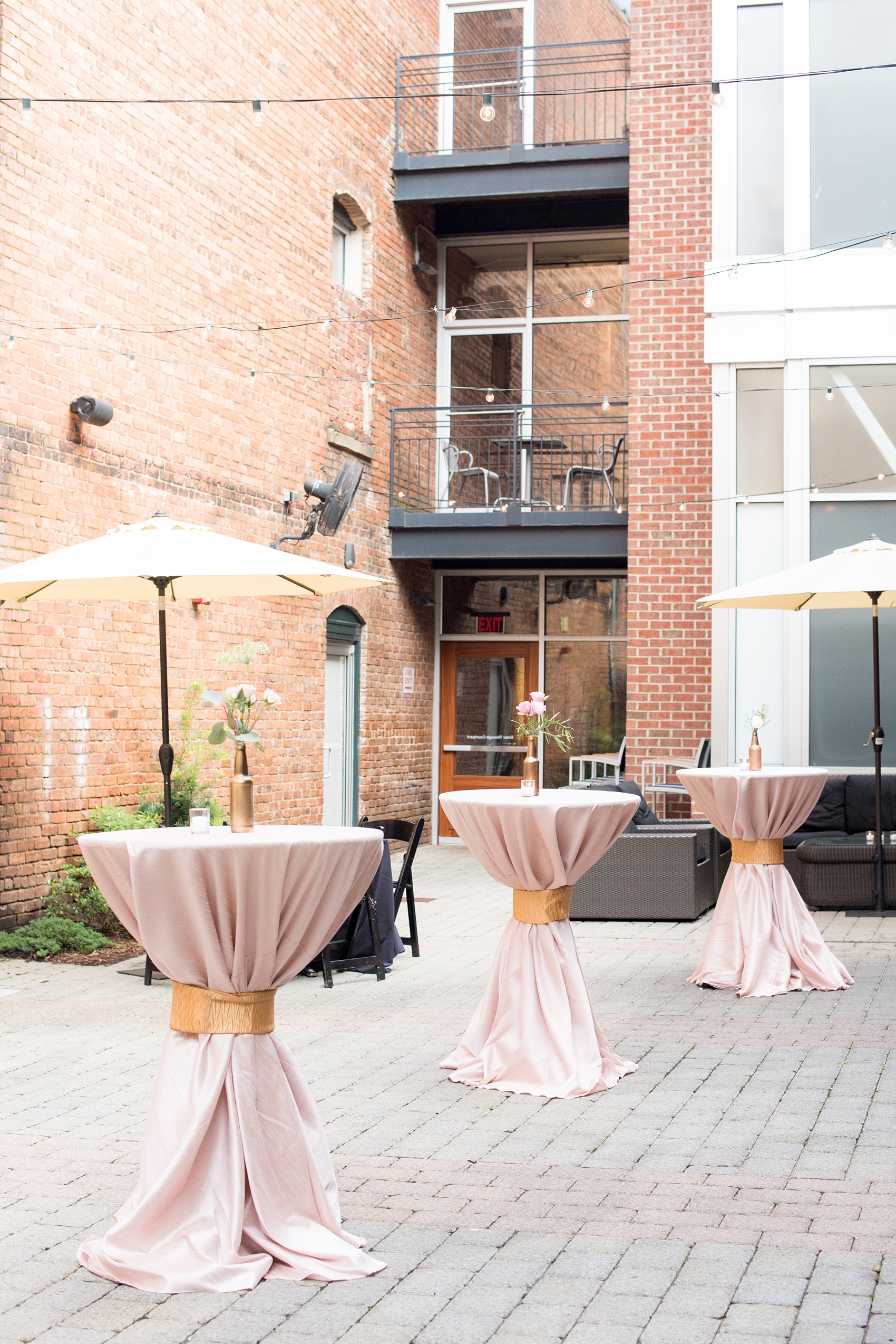 Mikkel Paige Photography photos from a downtown Raleigh wedding rehearsal dinner at Sitti restaurant. Picture of the pink hightop tables with blush linens and gold wraps. Centerpieces with gold bottles and flowers were on top of the tables.