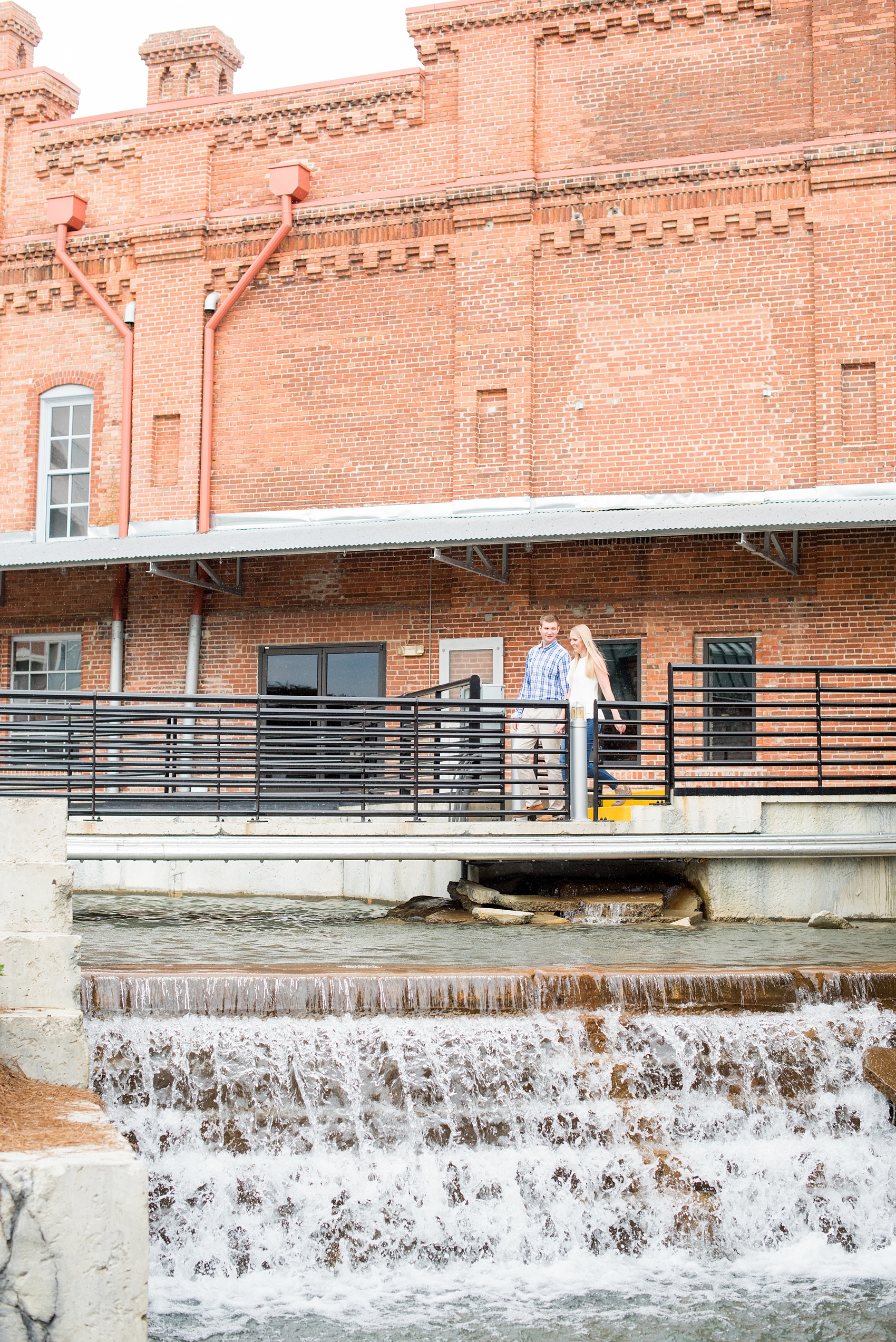 Mikkel Paige Photography photos from an engagement session at Durham's American Tobacco Campus in North Carolina. Picture of the couple walking over a bridge on the waterfalls.