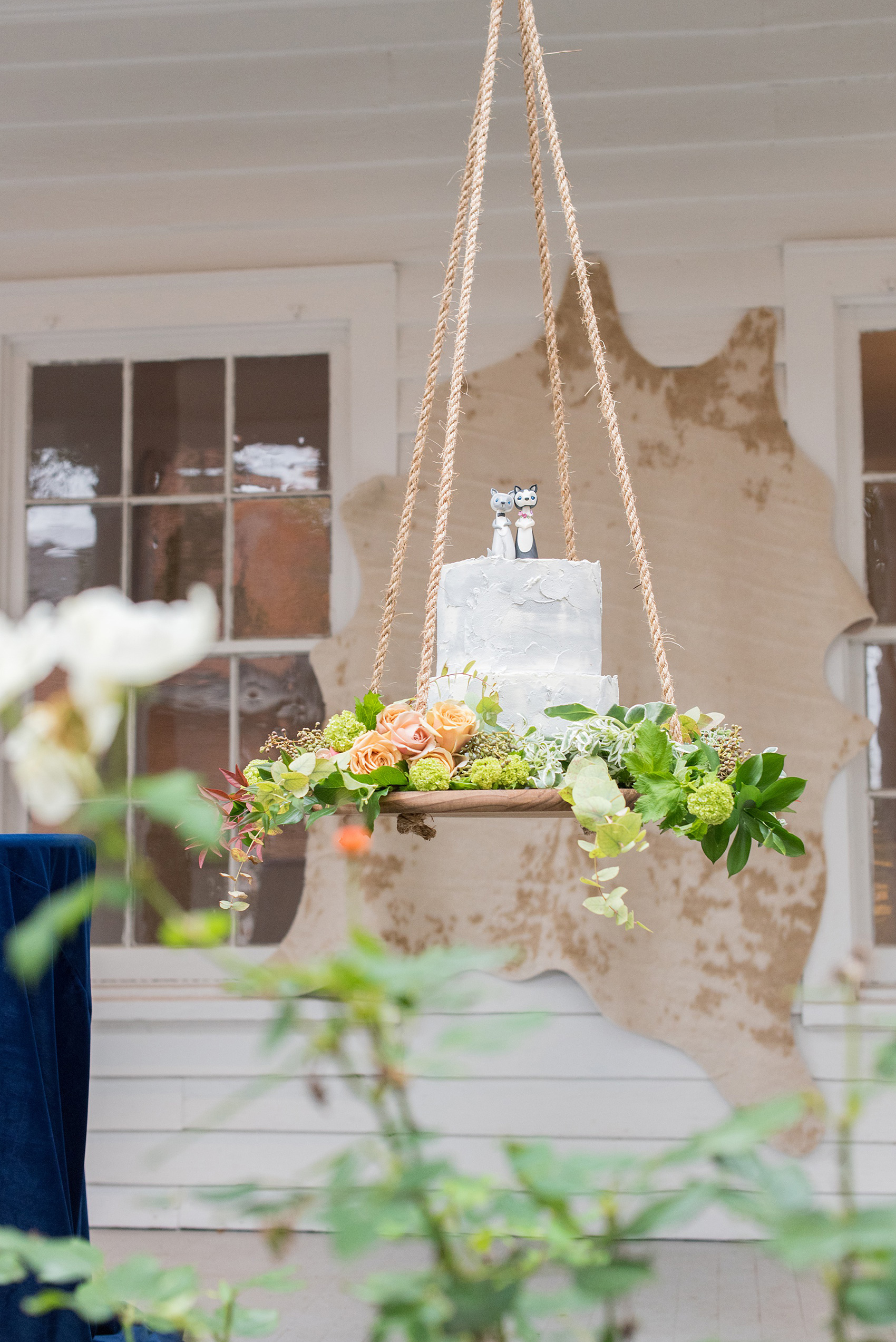 Mikkel Paige Photography photos from a wedding at Leslie-Alford Mims House in North Carolina. Picture of a unique boho cake display: a grey concrete buttercream cake with clay animal cake topper, suspended from the venue on a rope display adorned with flowers.