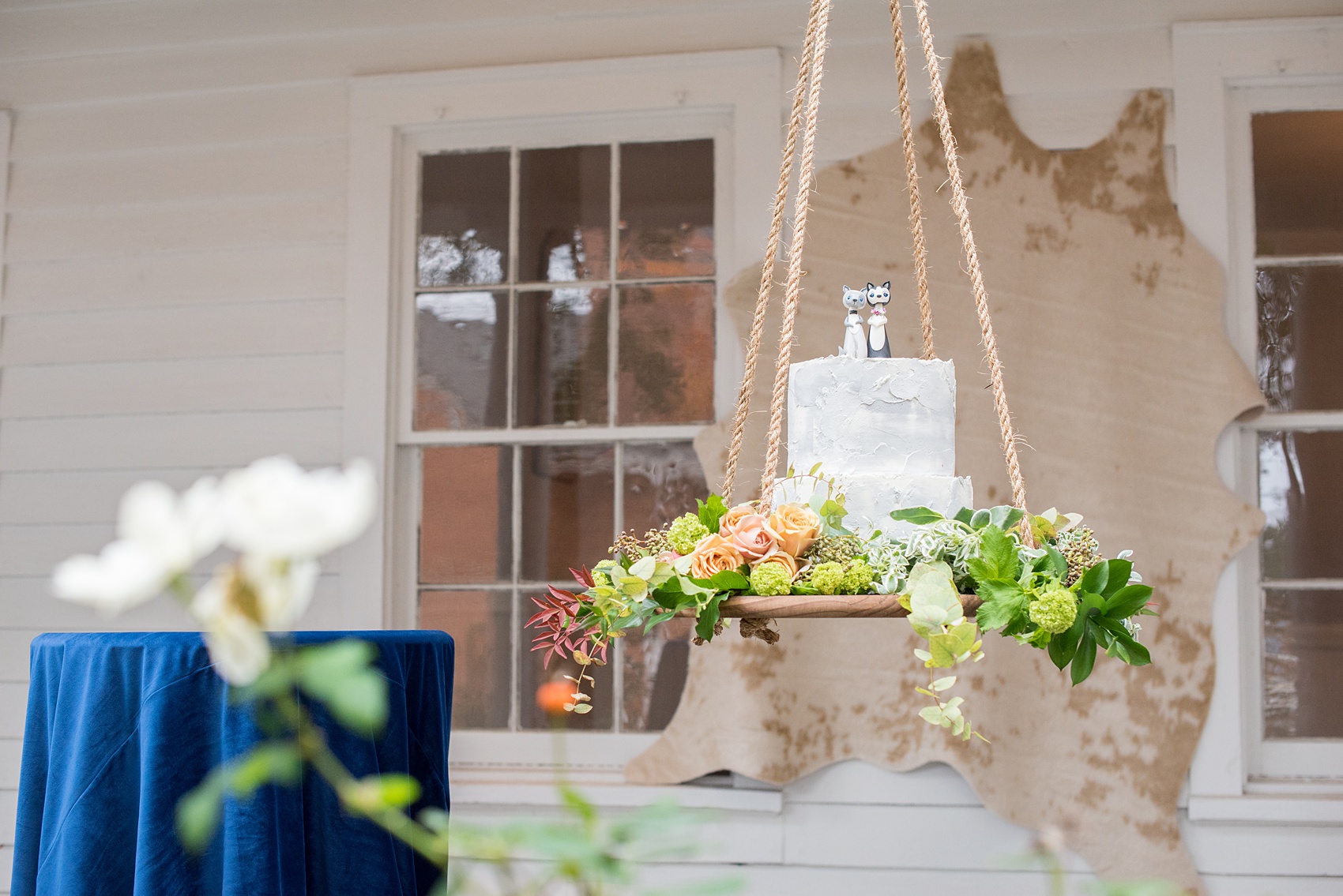 Mikkel Paige Photography photos from a wedding at Leslie-Alford Mims House in North Carolina. Picture of the concrete cake with clay animal cake topper, suspended from the venue on a rope display adorned with flowers.