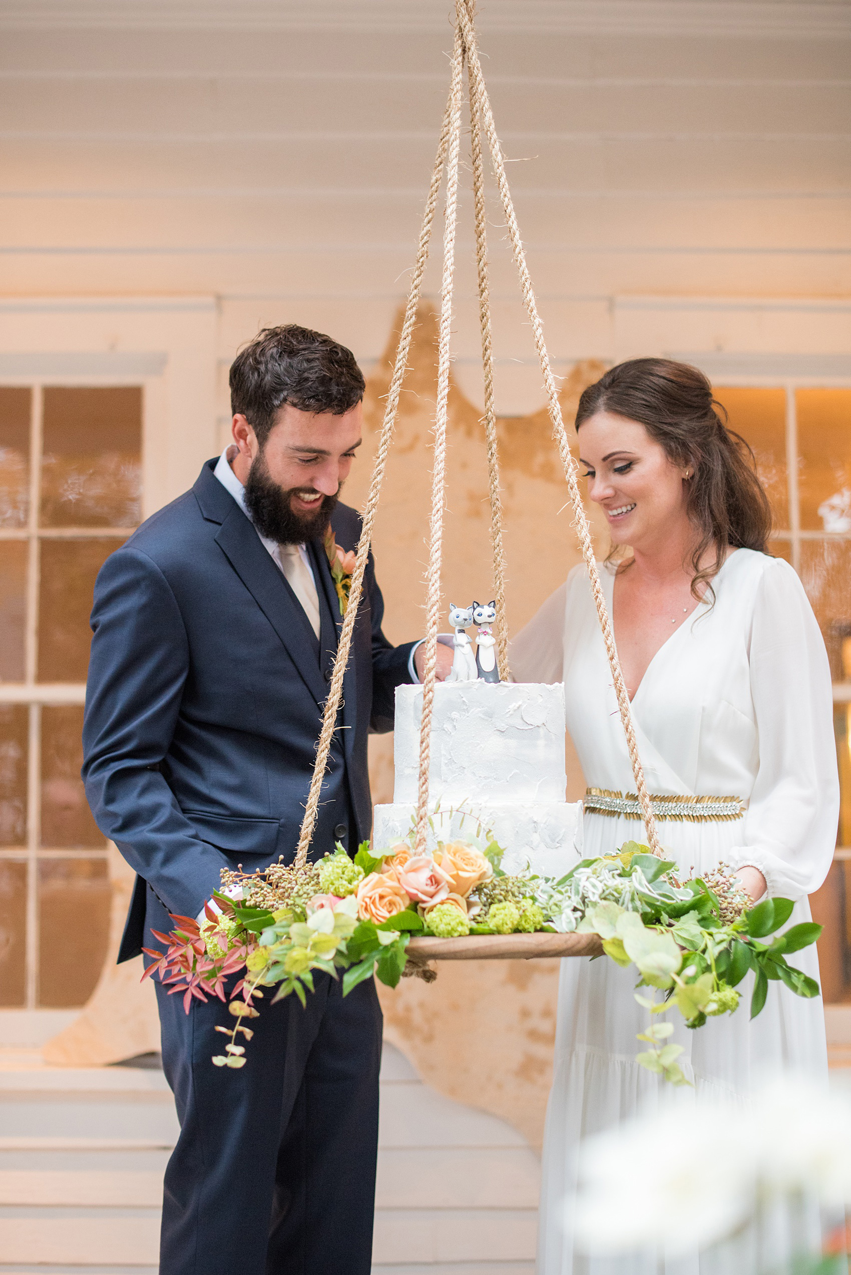 Mikkel Paige Photography photos from a wedding at Leslie-Alford Mims House in North Carolina. Picture of the bride and groom cutting their concrete cake with clay animal cake topper, suspended from the venue on a rope display adorned with flowers.