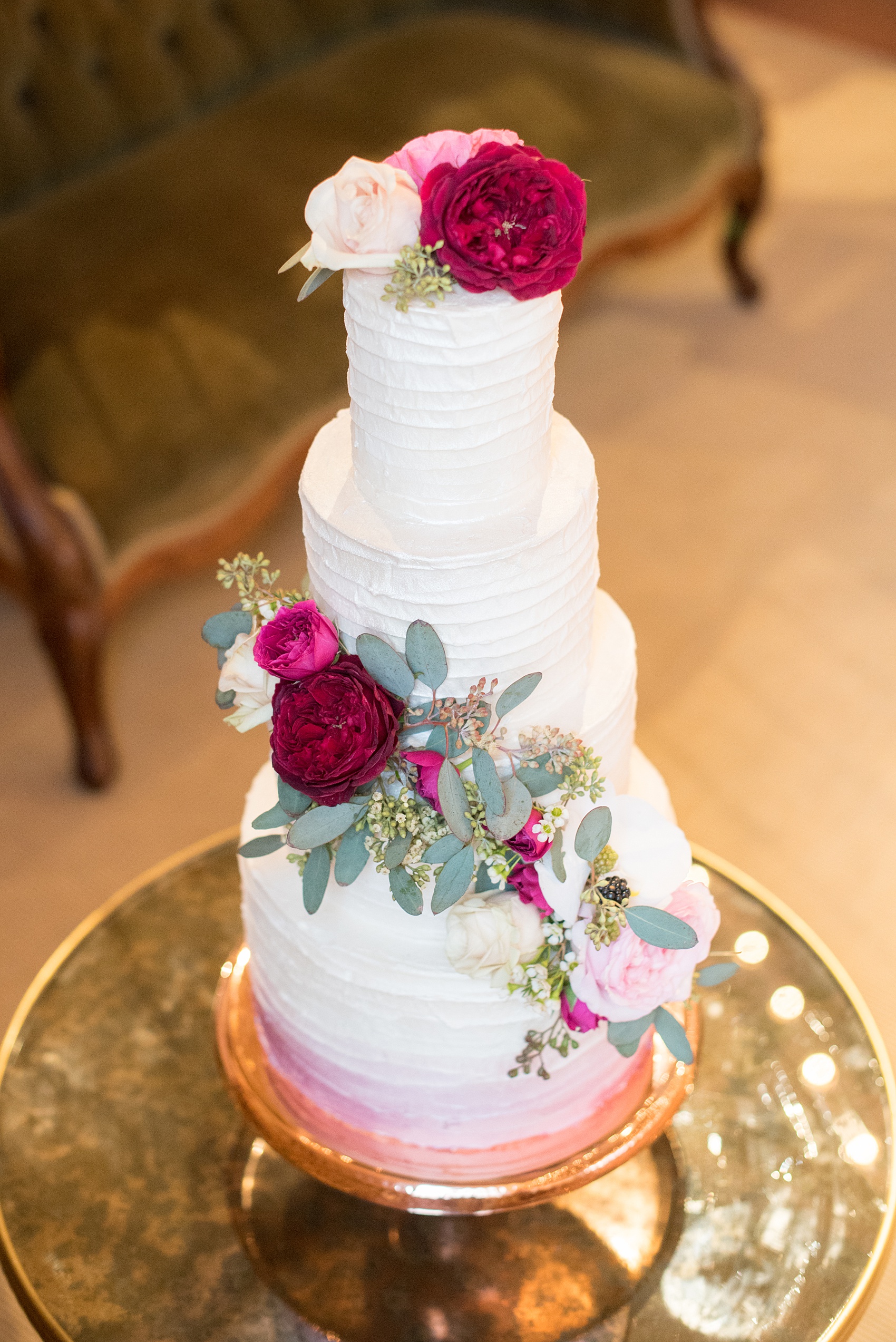 Photos from a wedding in downtown Raleigh, NC by Mikkel Paige Photography. The reception dessert options included cake and red and white macarons. The three tiered white buttercream creation was decorated with cascading fuchsia, maroon, burgundy and white flower and eucalyptus and dusted with luster powder in a pink ombre, by The Cupcake Shoppe.