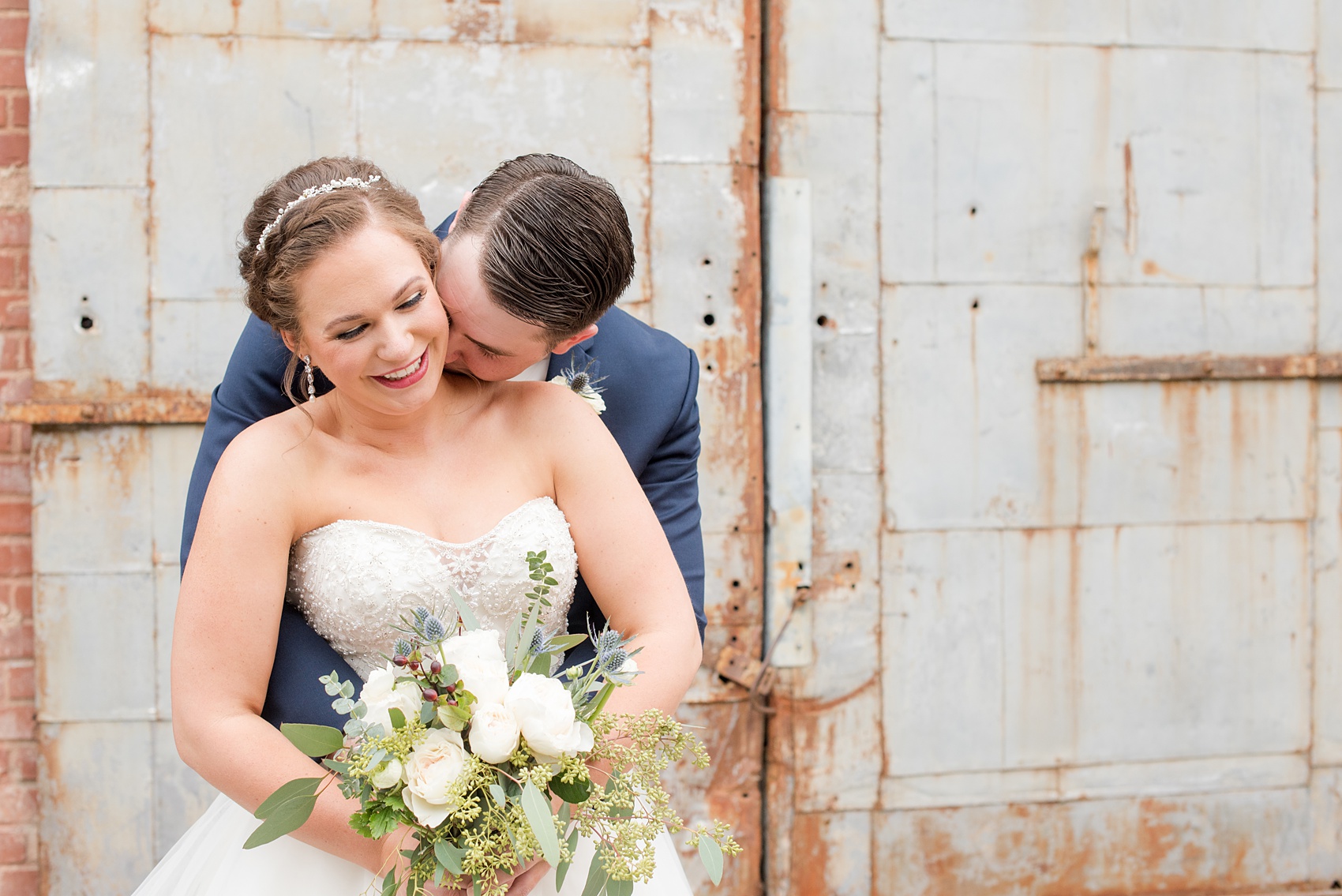 Durham wedding photos at The Cookery by Mikkel Paige Photography in North Carolina. In this cute picture of the bride and groom he kisses her neck in front of the venue's rustic metal door.