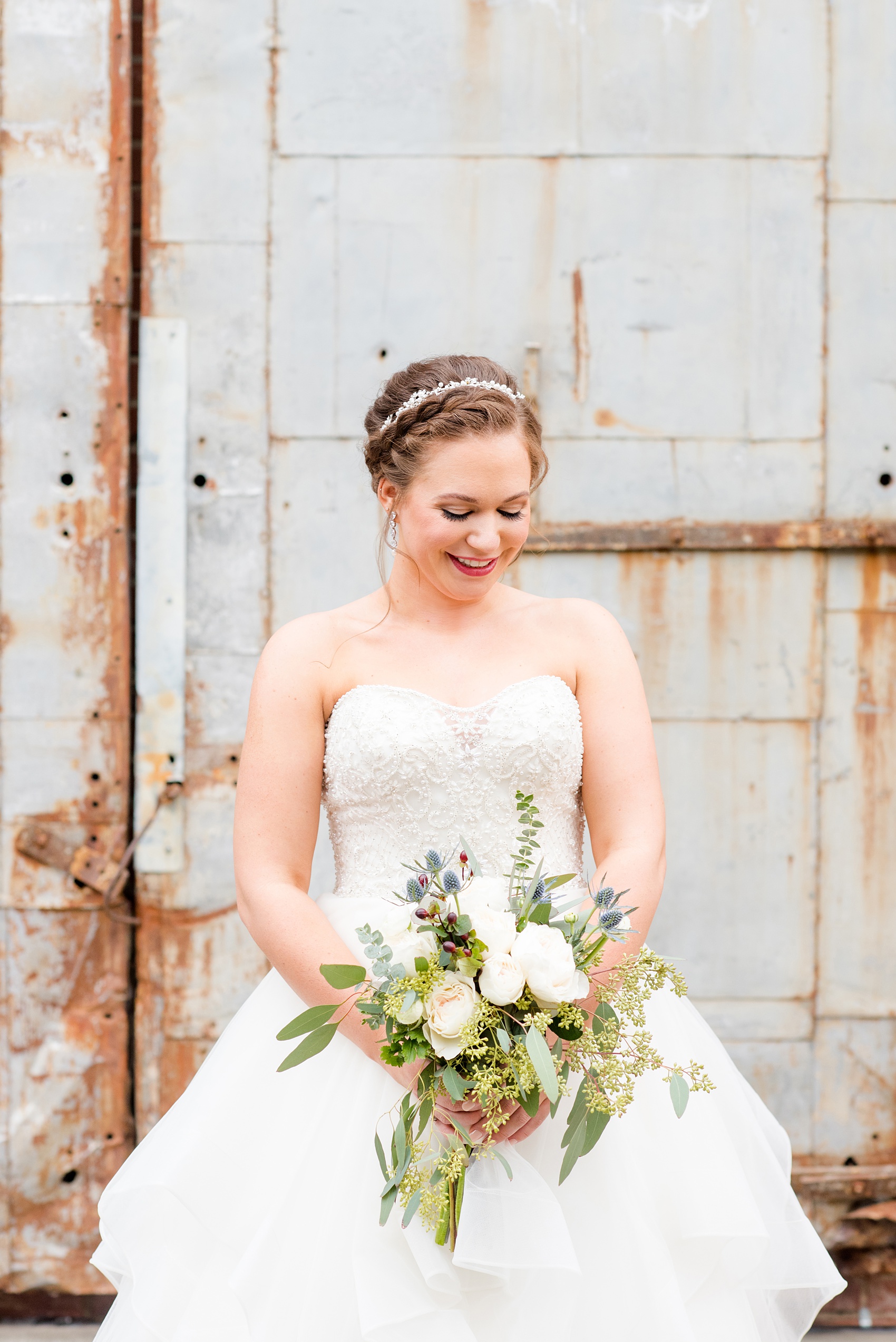 Durham wedding photos at The Cookery by Mikkel Paige Photography in North Carolina. The bride wore a strapless beaded gown, braided up-do and held a bouquet of roses, blue thistle and eucalyptus for her winter day. In this picture she's posing in front of the venue's rustic doors.