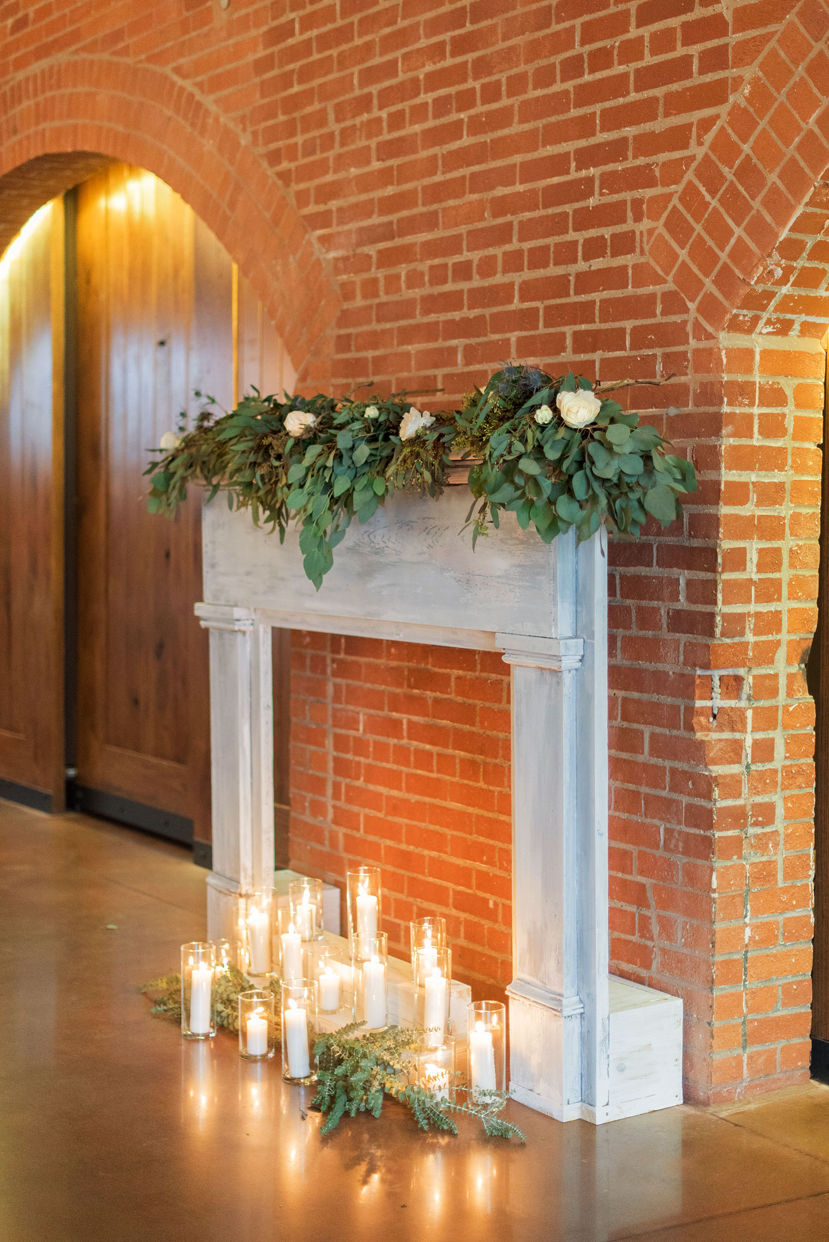 Durham wedding photos at The Cookery by Mikkel Paige Photography in North Carolina. The bride's father created this faux fireplace, decorated with lots of candlelight and green garland and white roses. It also served as the indoor ceremony backdrop.