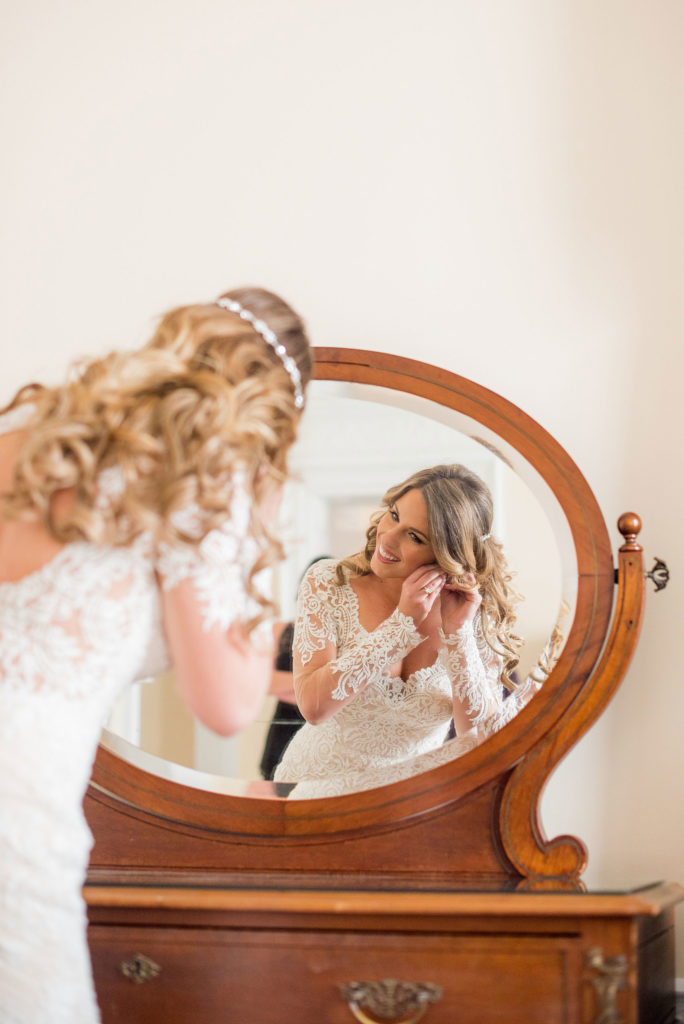 Wedding photos at Sleepy Hollow Country Club for a winter reception in January by Mikkel Paige Photography. A picture of the bride looking into the mirror to put on her vintage diamond and pearl earrings.