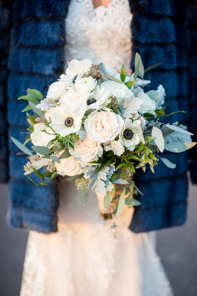 Wedding photos at Sleepy Hollow Country Club for a winter reception in January by Mikkel Paige Photography. A detail picture of the bride's winter bouquet, with white and green flowers, including anemones, eucalyptus and roses against her blue fur coat.