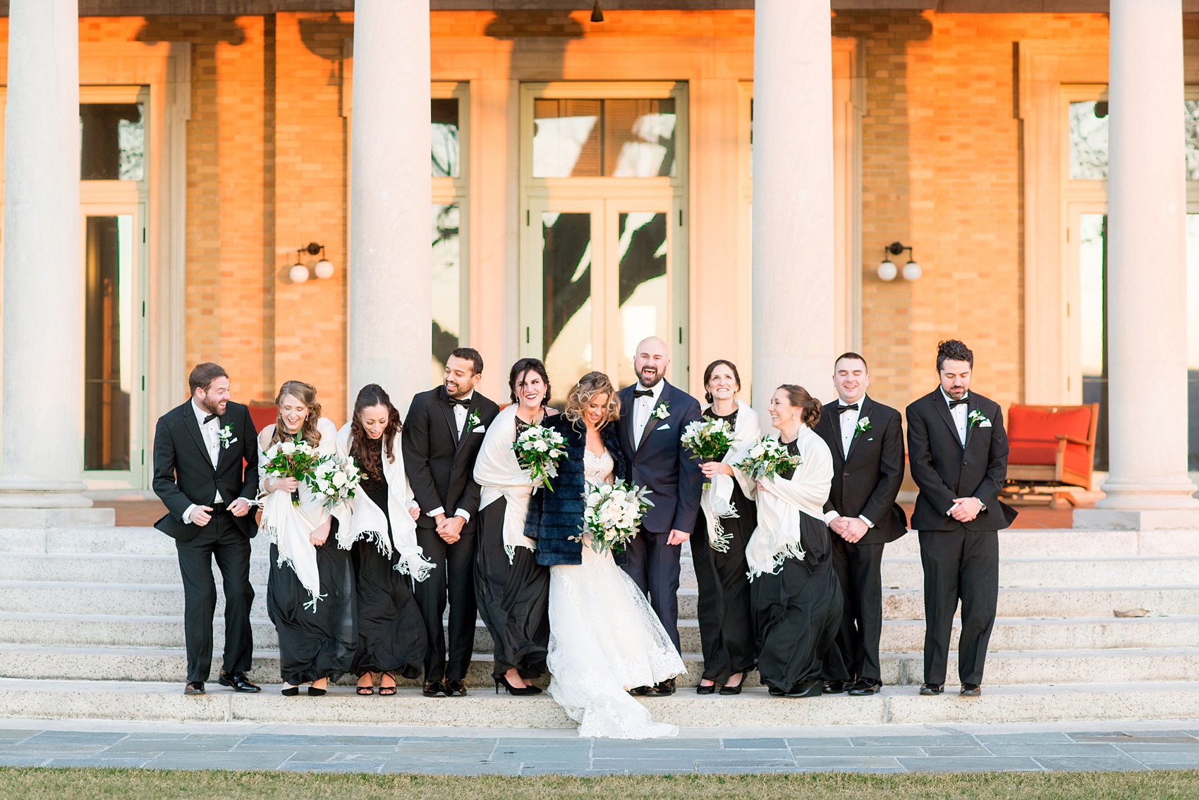 Wedding photos at Sleepy Hollow Country Club for a winter reception in January by Mikkel Paige Photography. The bridal party is pictured with white shawls, black lace gowns, and black tuxedos on the iconic stairs with columns of this New York venue.