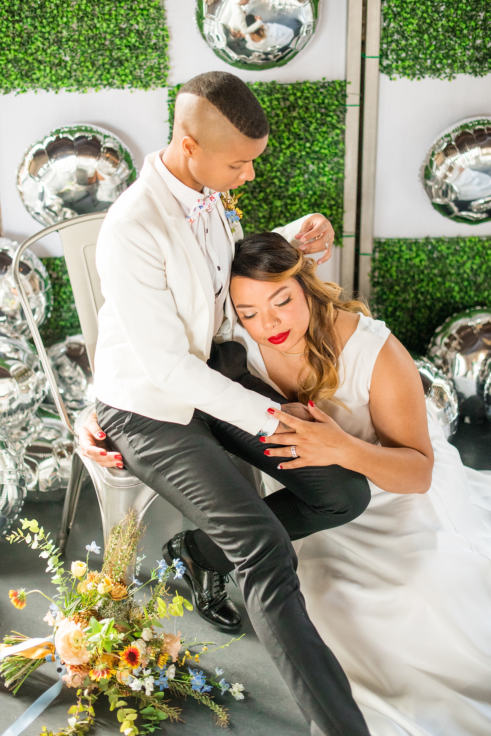 Mikkel Paige Photography photos of a wedding at the Green Building in Brooklyn, New York. This same sex, gay marriage styled shoot was created by Color Pop Events. They recited vows during their ceremony in front of silver mylar balloons and green square boxwood patches. The bride wore a white, silk cowl neck gown and the bride groom in a white coat with colorful bow tie.