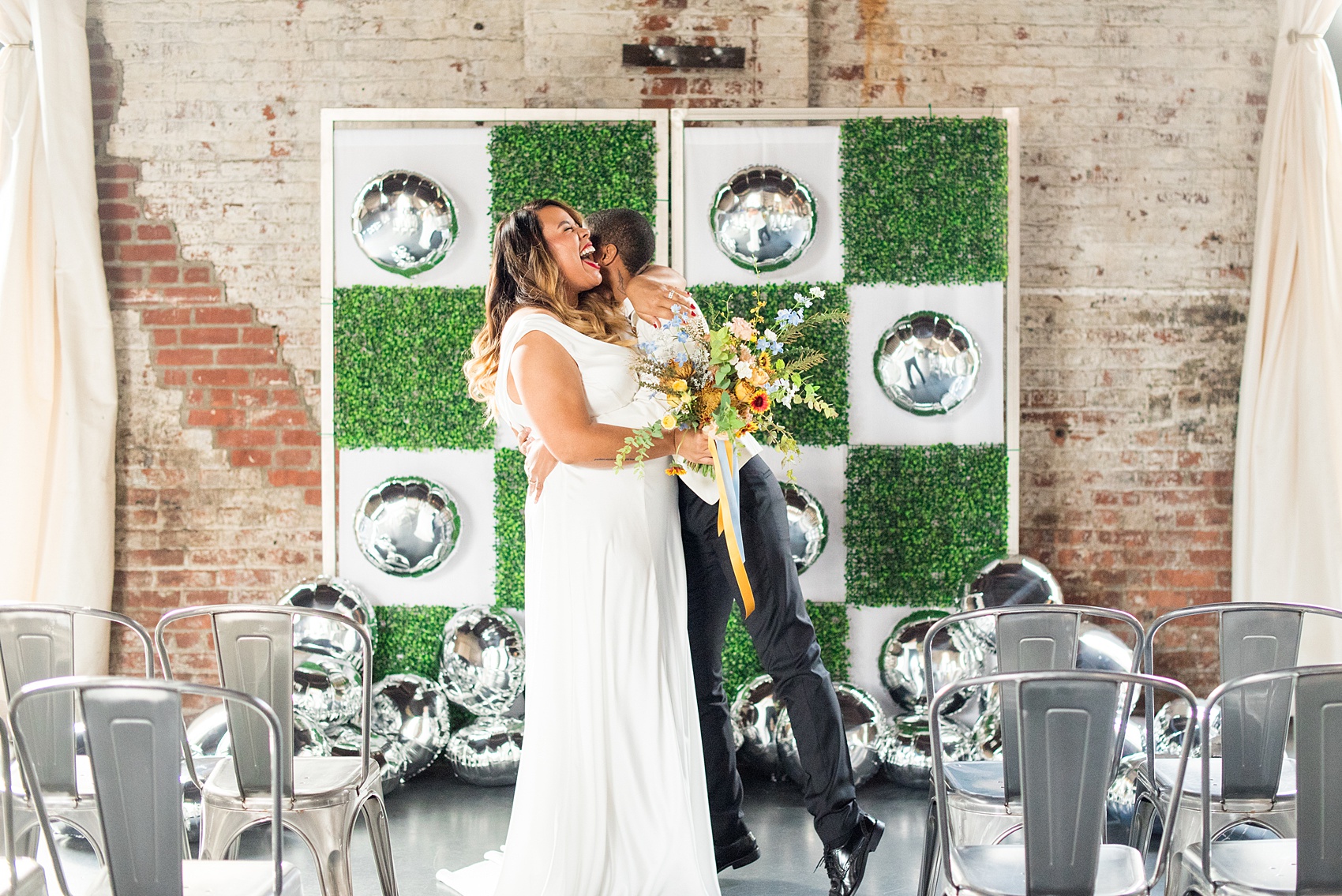 Mikkel Paige Photography photos of a wedding at the Green Building in Brooklyn, New York. This same sex, gay marriage styled shoot was created by Color Pop Events. They recited vows during their ceremony in front of silver mylar balloons and green square boxwood patches. The bride wore a white, silk cowl neck gown and the bride groom in a white coat with colorful bow tie.