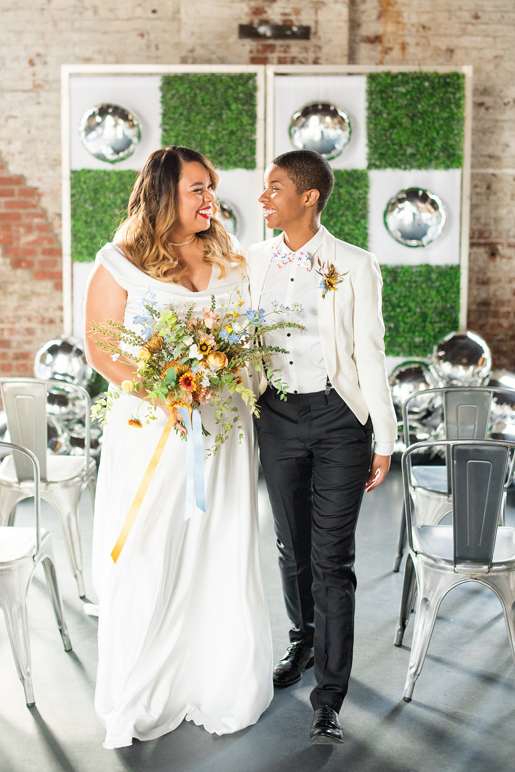 Mikkel Paige Photography photos of a wedding at the Green Building in Brooklyn, New York. This same sex, gay marriage styled shoot was created by Color Pop Events. They recited vows during their ceremony in front of silver mylar balloons and green square boxwood patches. The bride wore a white, silk cowl neck gown and the bride groom in a white coat with colorful bow tie.