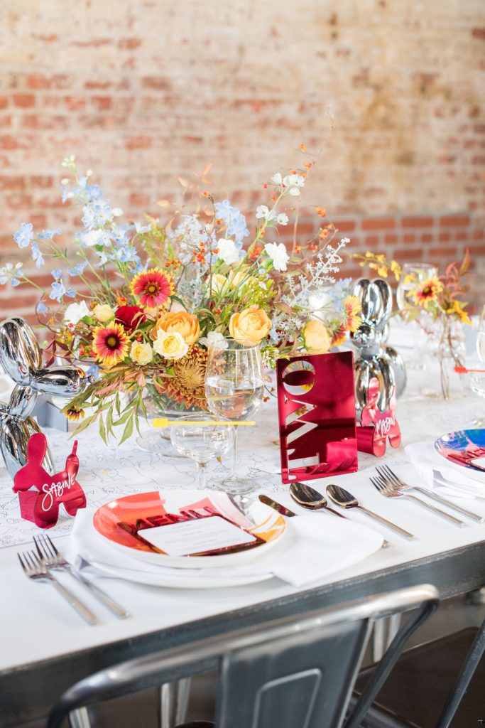 Mikkel Paige Photography images of a wedding at the Green Building in Brooklyn, New York. This same sex, gay marriage styled shoot was created by Color Pop Events. The urban dinner table was styled by Taylor and Hov, inspired by Jeff Koons and neo pop art. Flower centerpieces are by Abby Tabak Studio. The red mirror laser cut table number was propped up on a coloring book table runner.