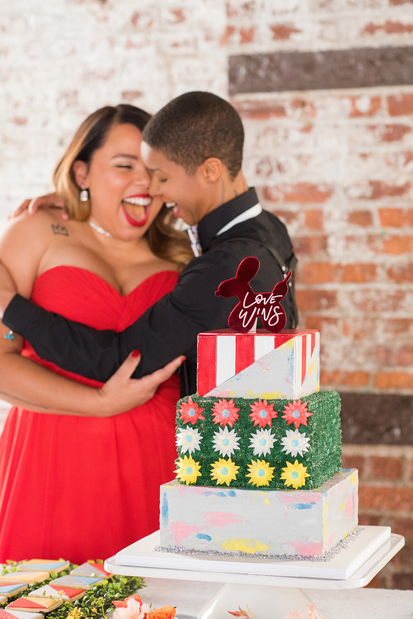 Mikkel Paige Photography images of a wedding at the Green Building in Brooklyn, New York. This same sex, gay marriage styled shoot was created by Color Pop Events. The couple wore second red, black and silver outfits. The cake was finished with a custom balloon animal, mirror red "Love Wins" topper and had three square tiers with red, white and yellow elements dashed with green piping.