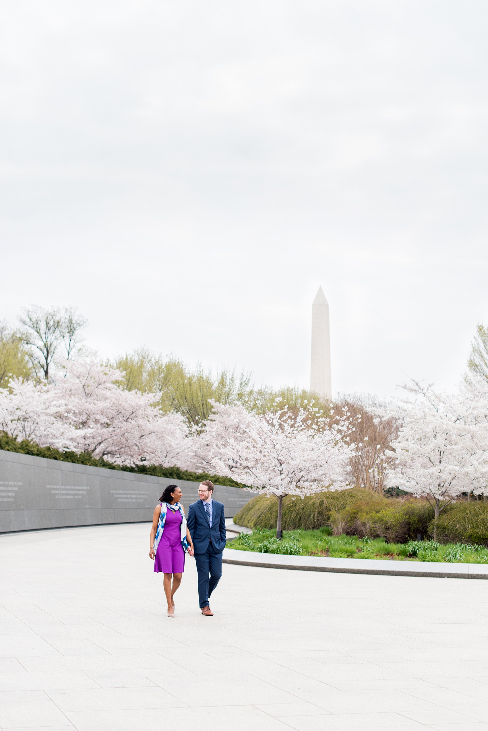 DC Cherry Blossoms Engagement Photos by Mikkel Paige Photography. Spring flowers at the Martin Luther King Jr. memorial with the Washington Monument in the background, at the nation's Capitol with an interracial couple in colorful outfits. #DCCherryBlossoms #CherryBlossoms #EngagementPhotos #SpringEngagementPhotos #CherryBlossomsEngagementPhotos